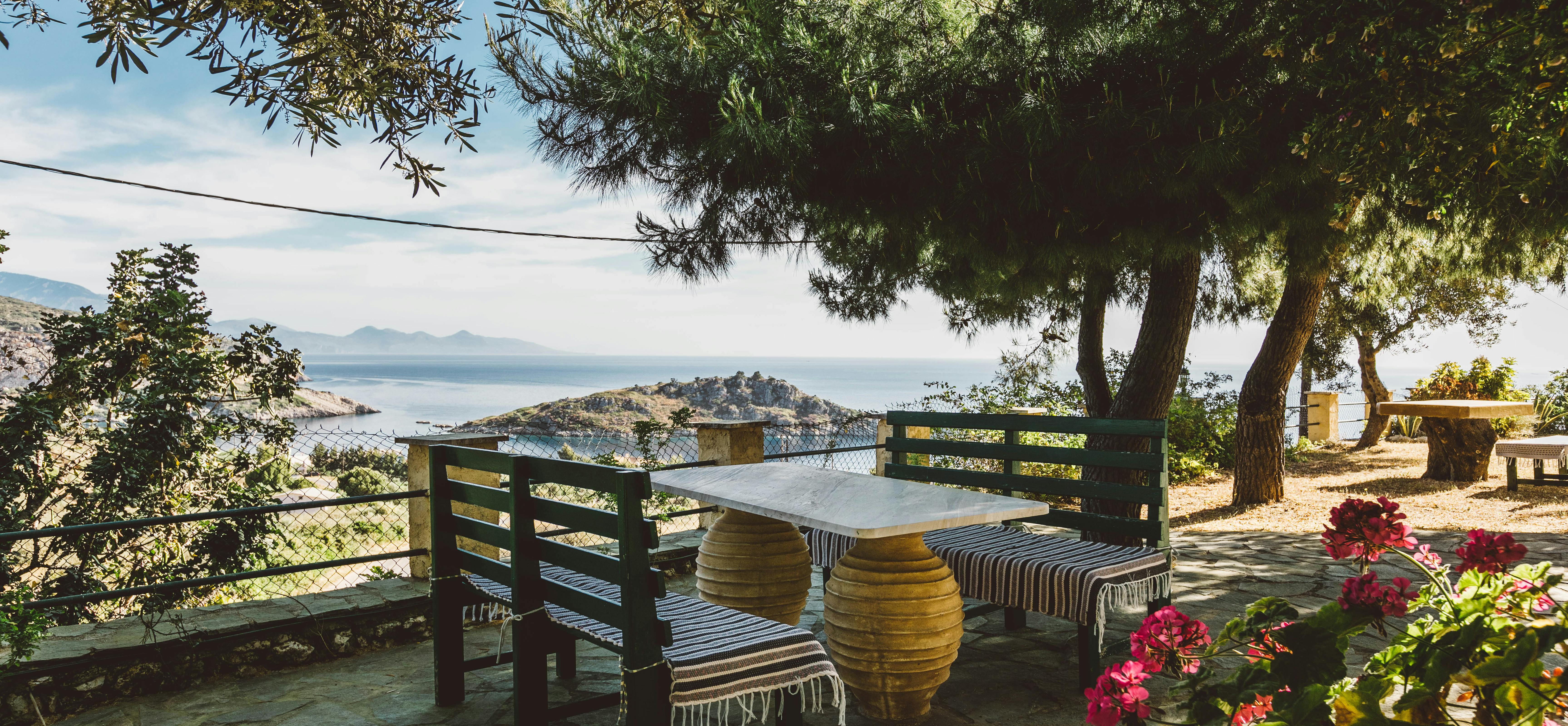 Shaded terrace seating area with rustic wooden furniture, striped textiles, and panoramic coastal views framed by olive trees.