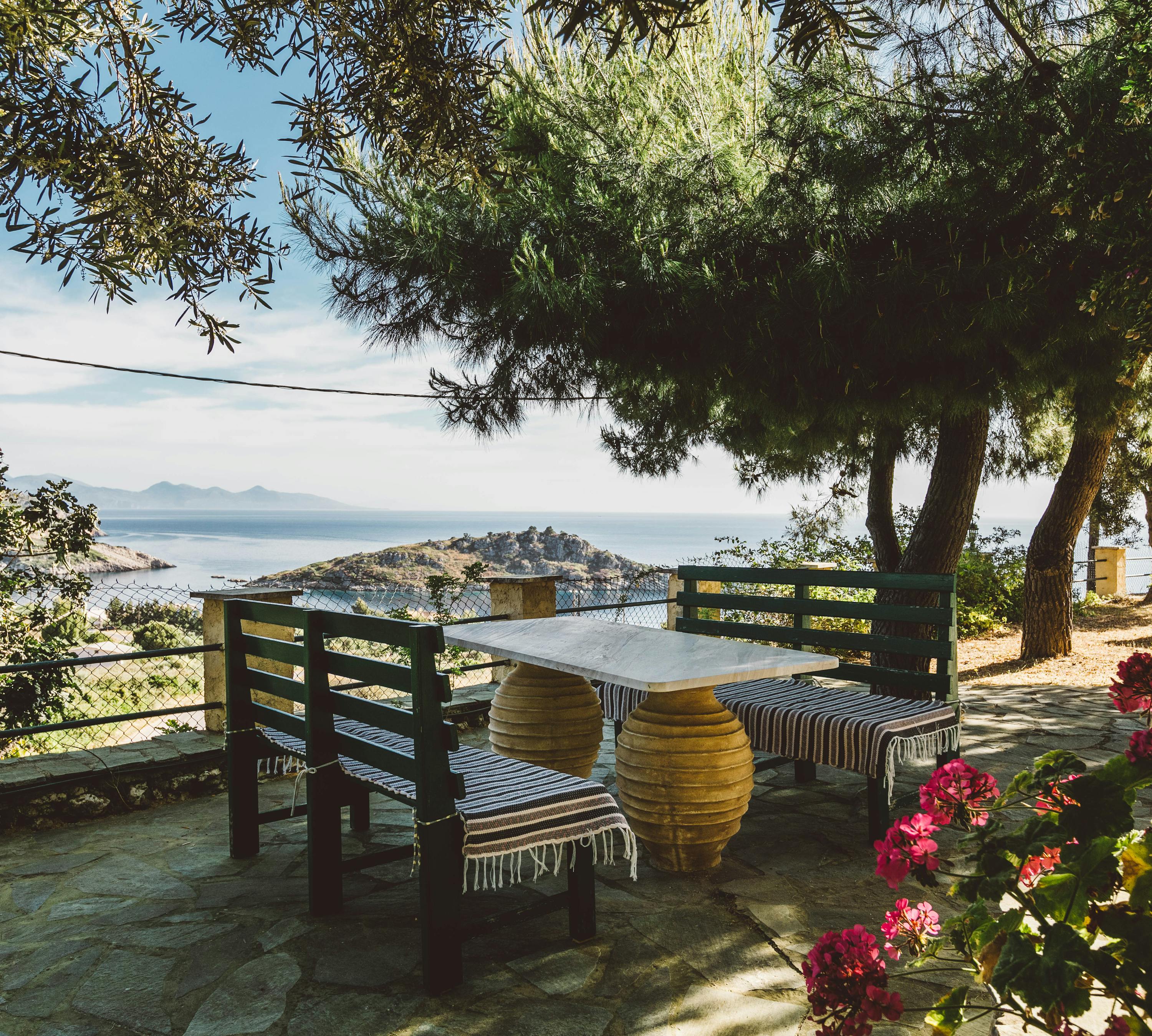 Shaded terrace seating area with rustic wooden furniture, striped textiles, and panoramic coastal views framed by olive trees.