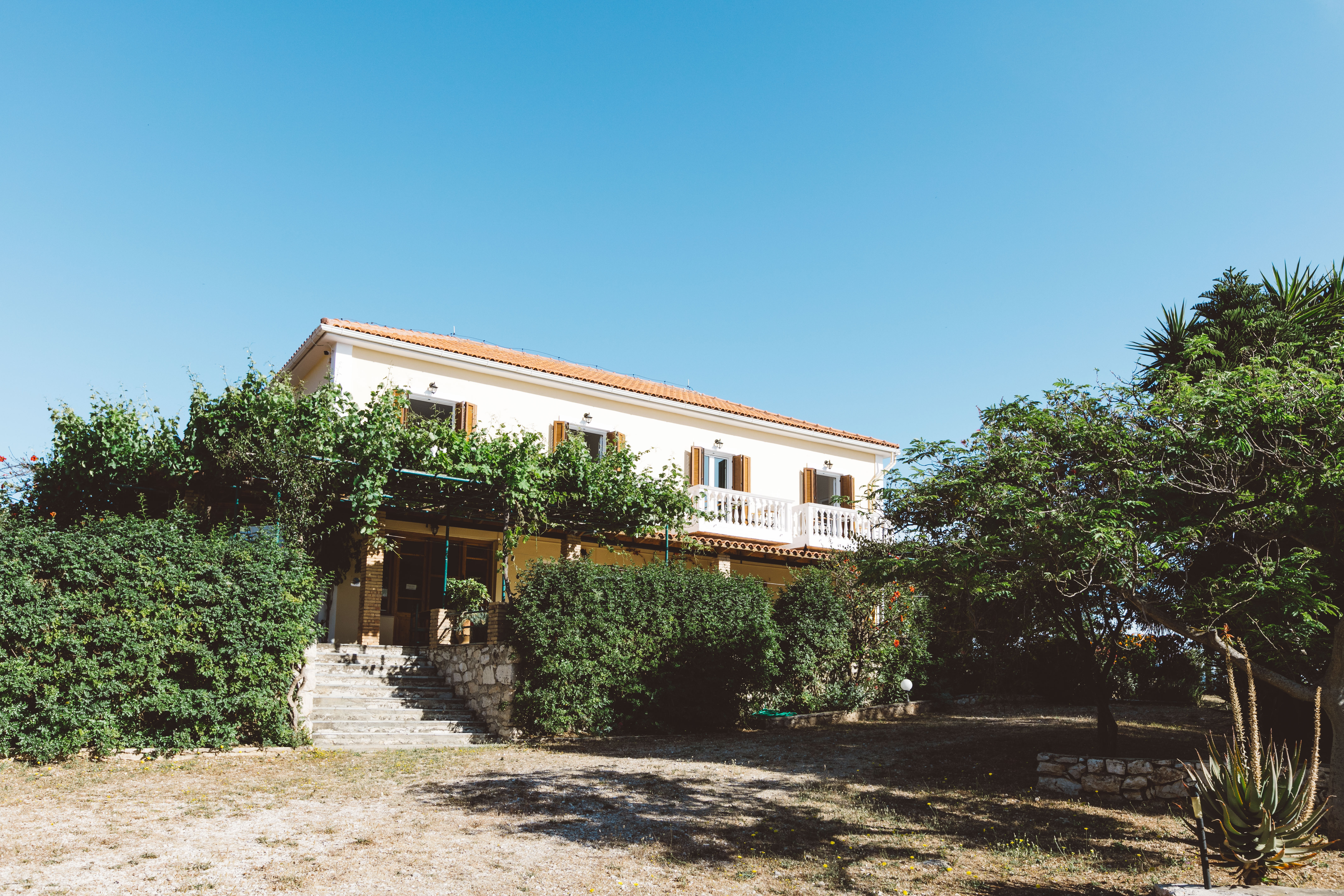 Two-story Mediterranean villa with terracotta roof, white balustrade balcony, and lush garden surroundings under clear blue sky.