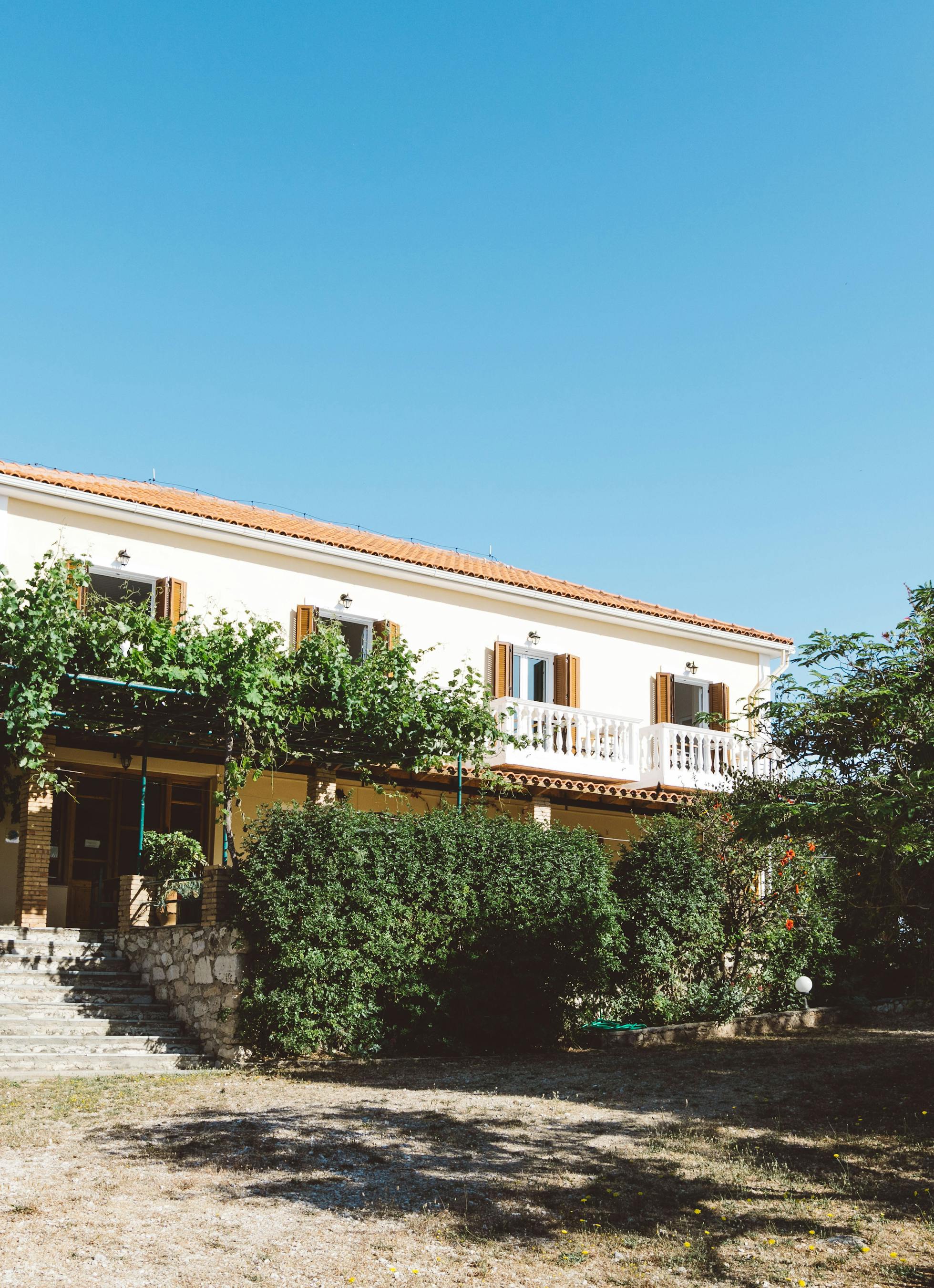 Two-story Mediterranean villa with terracotta roof, white balustrade balcony, and lush garden surroundings under clear blue sky.