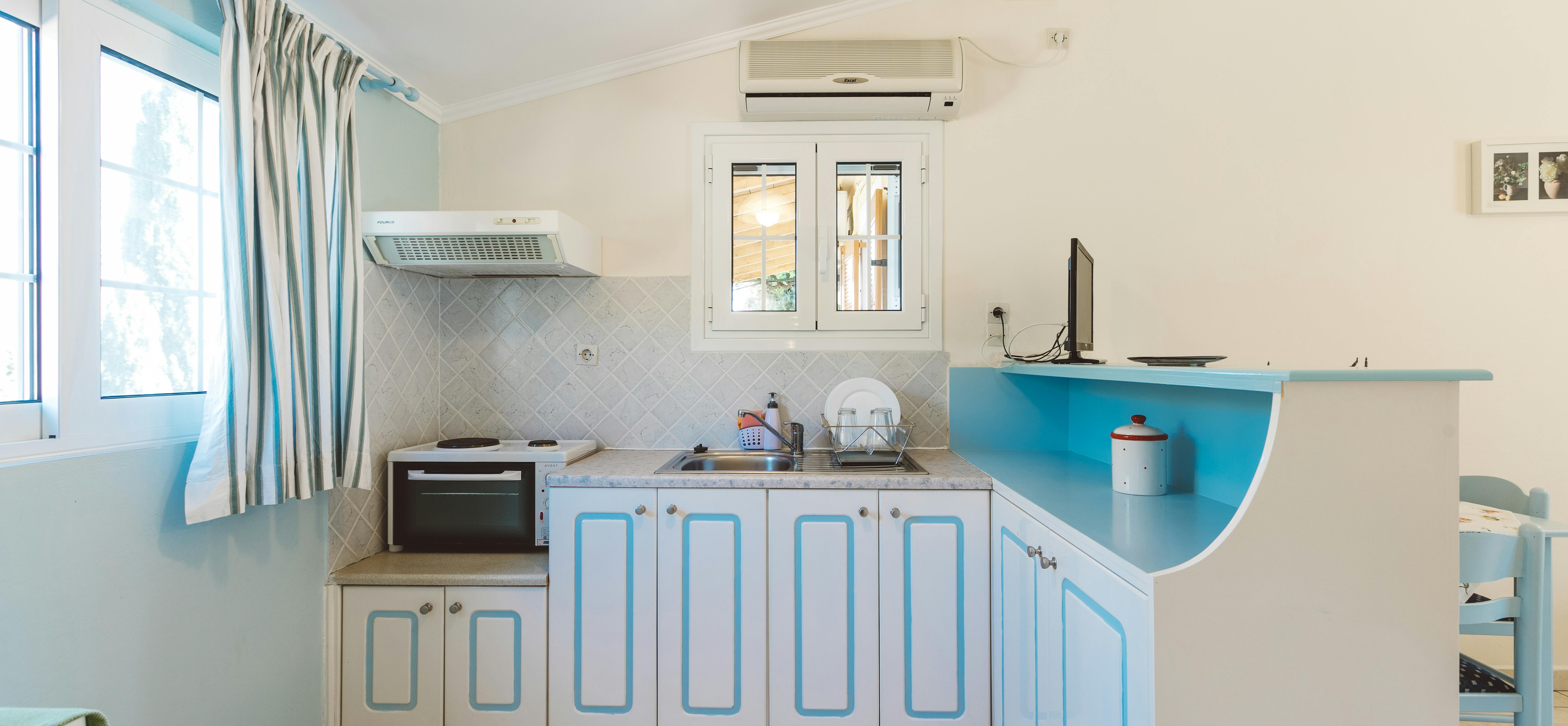 Compact kitchenette with white cabinets trimmed in turquoise, small appliances, and air conditioning unit.