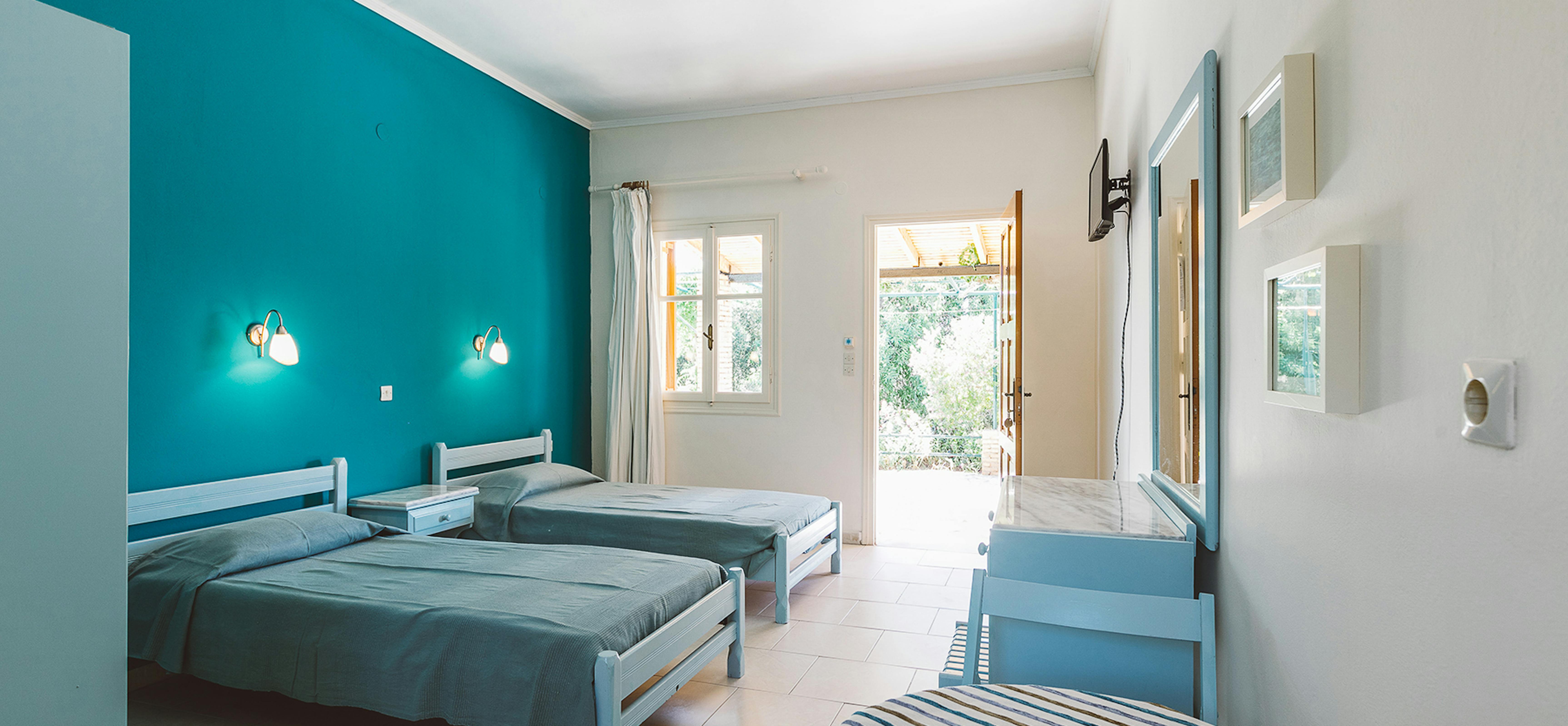 Ground-floor bedroom with turquoise accent wall, three twin beds with striped linens, and doors opening to garden.