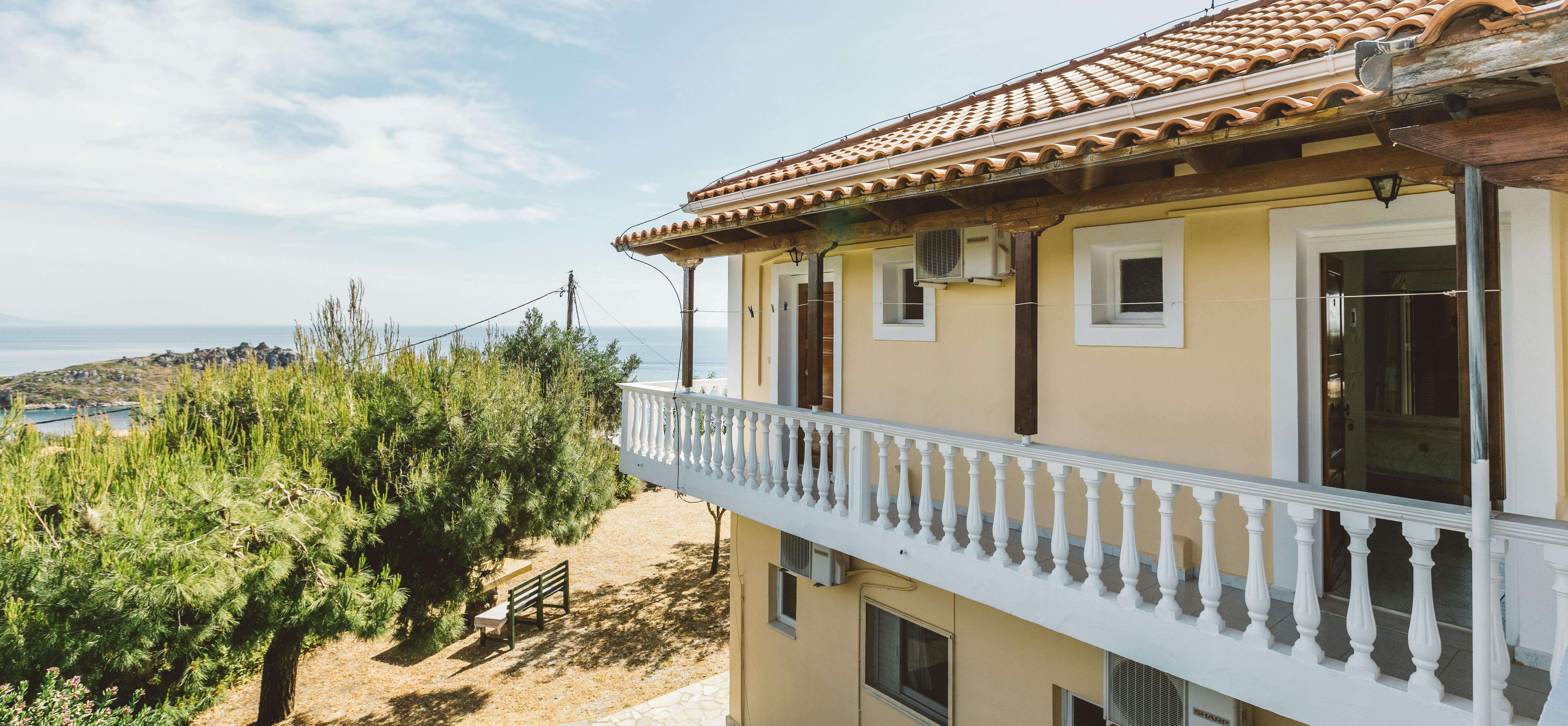 Upper balcony exterior showing terracotta roof overhang, white balustrade, and sea views through vegetation.