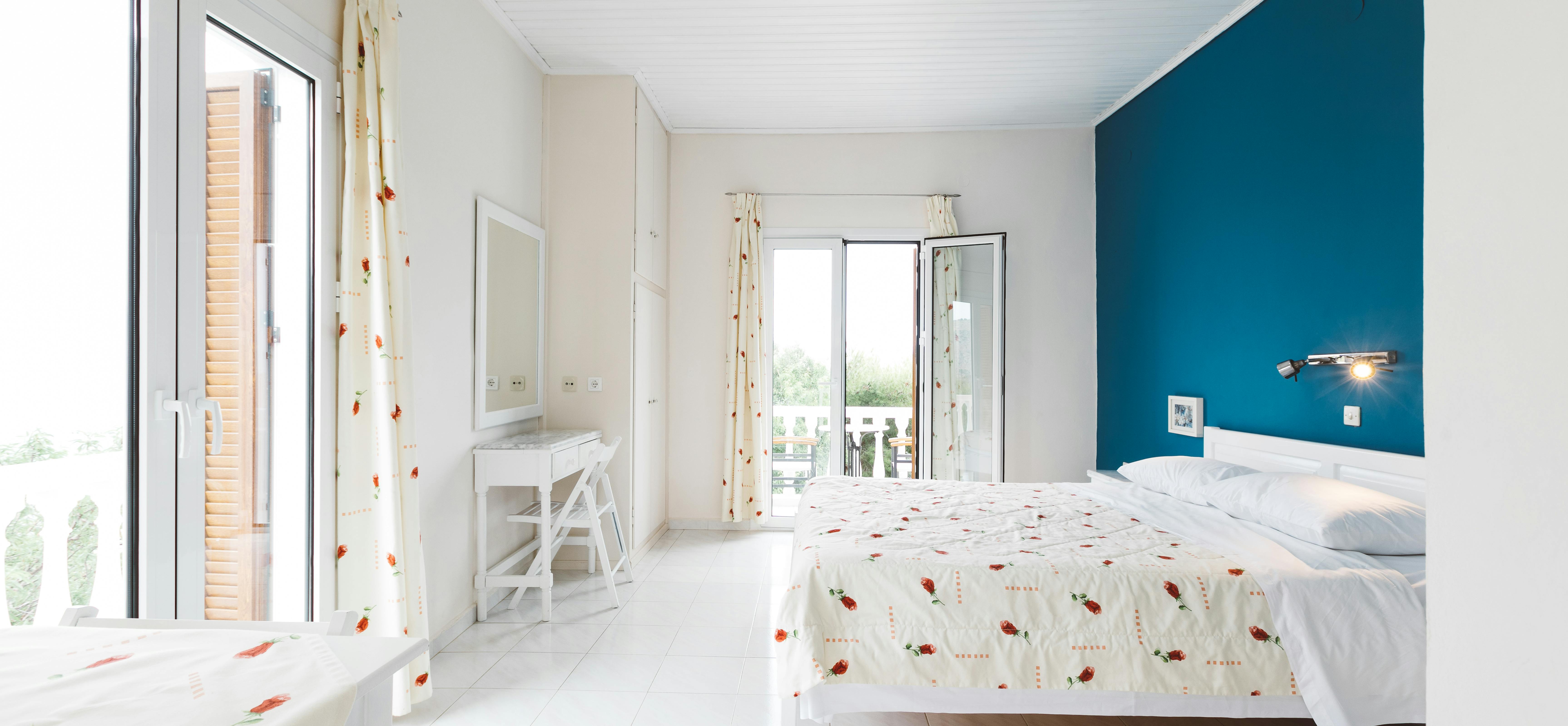 Bright bedroom with turquoise accent wall, white furnishings, balcony doors, and poppy-patterned linens and curtains.