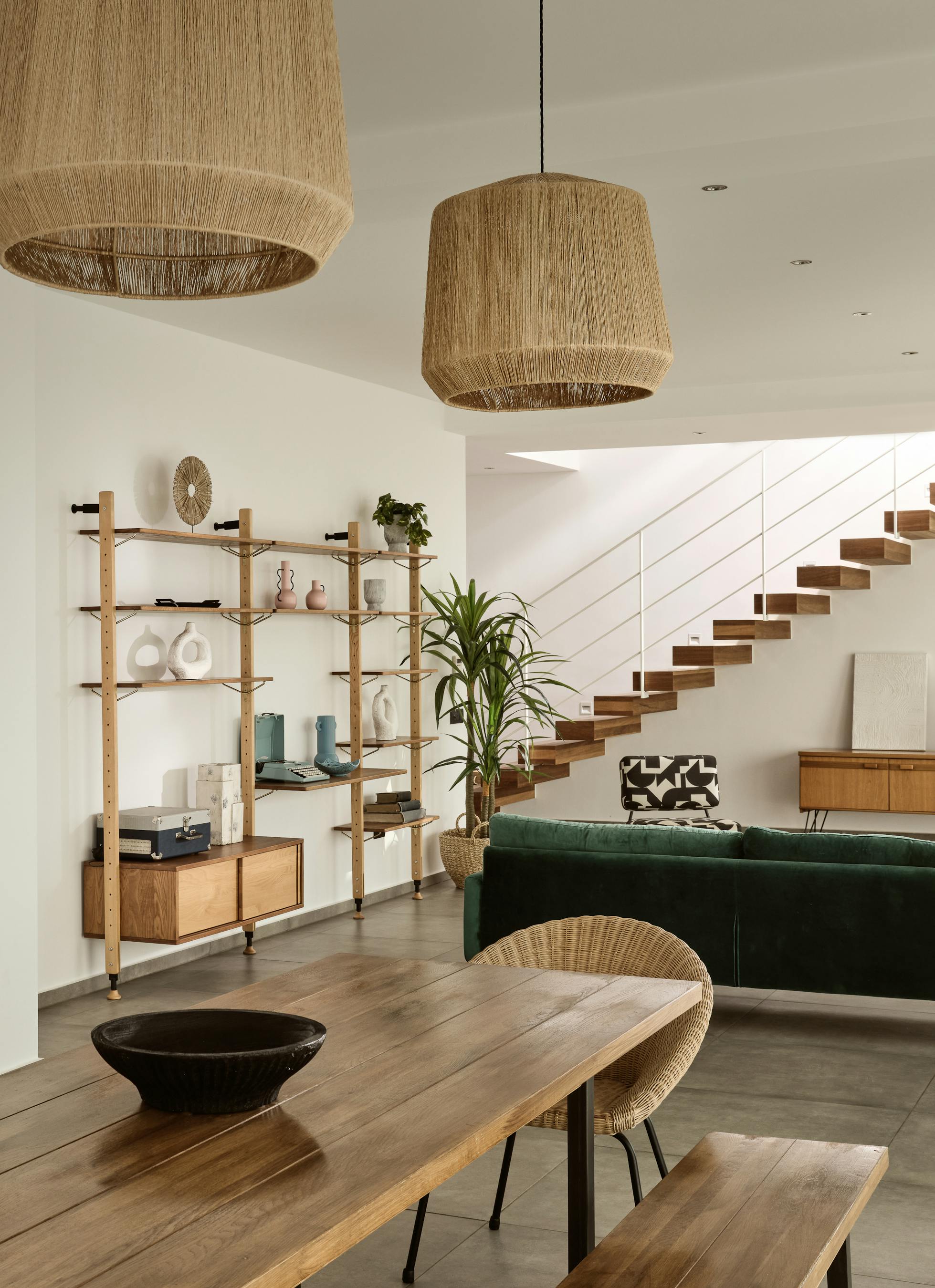 Open-plan living and dining area with wooden table, green velvet sofa, floating staircase, and natural pendant lighting.