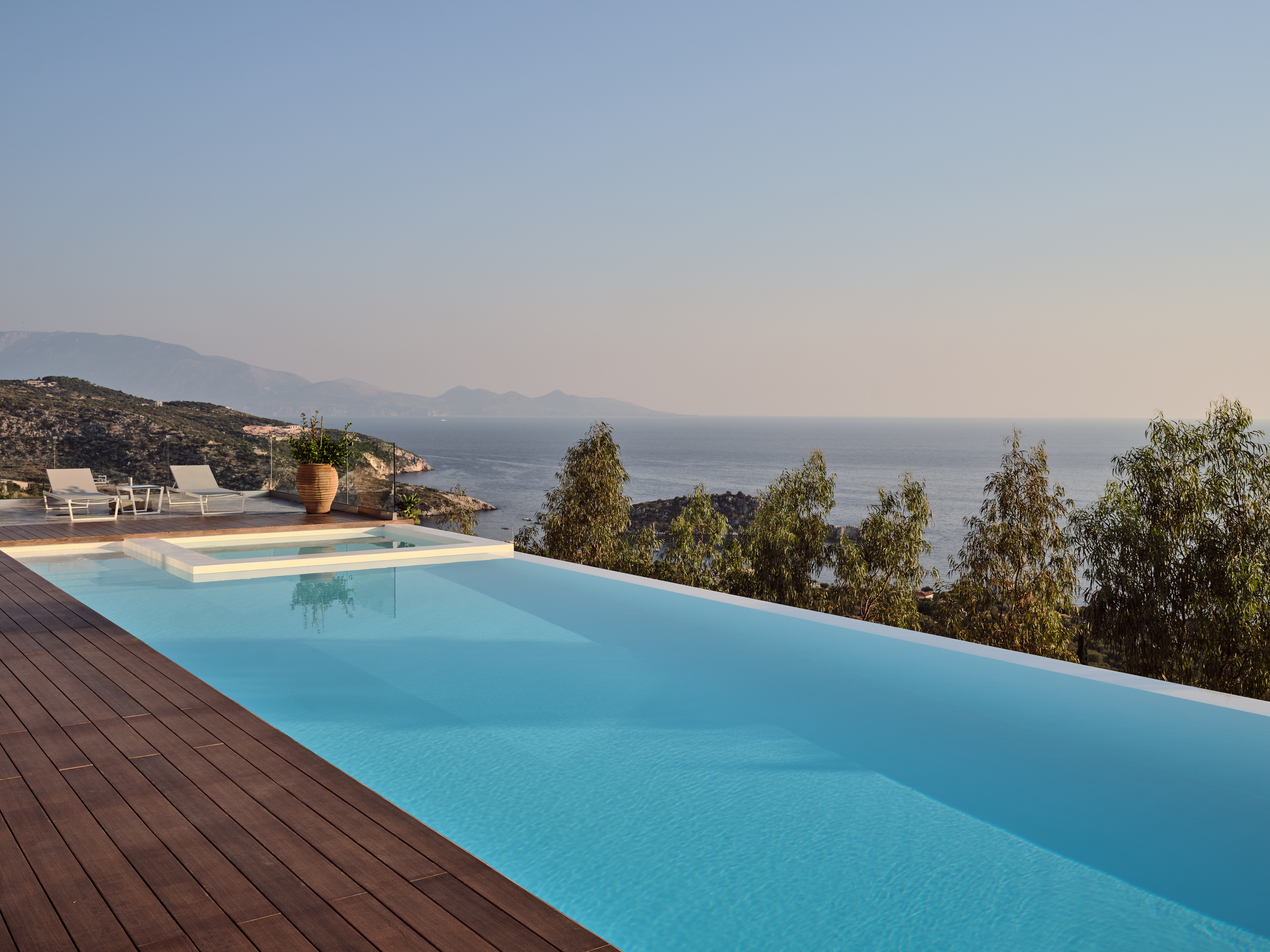 Infinity pool with wooden deck overlooking misty coastal landscape and distant mountains at dusk.