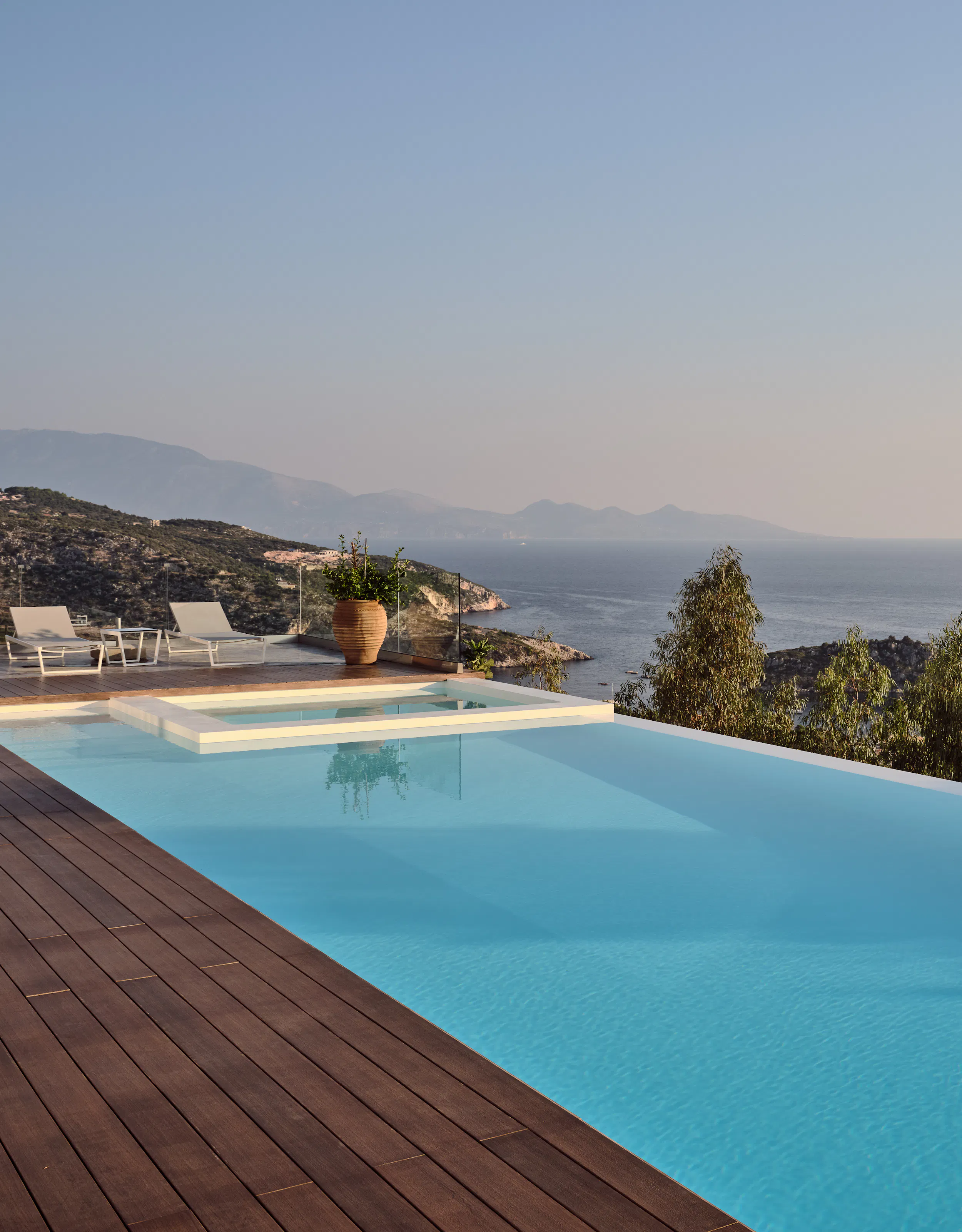 Infinity pool with wooden deck overlooking misty coastal landscape and distant mountains at dusk.