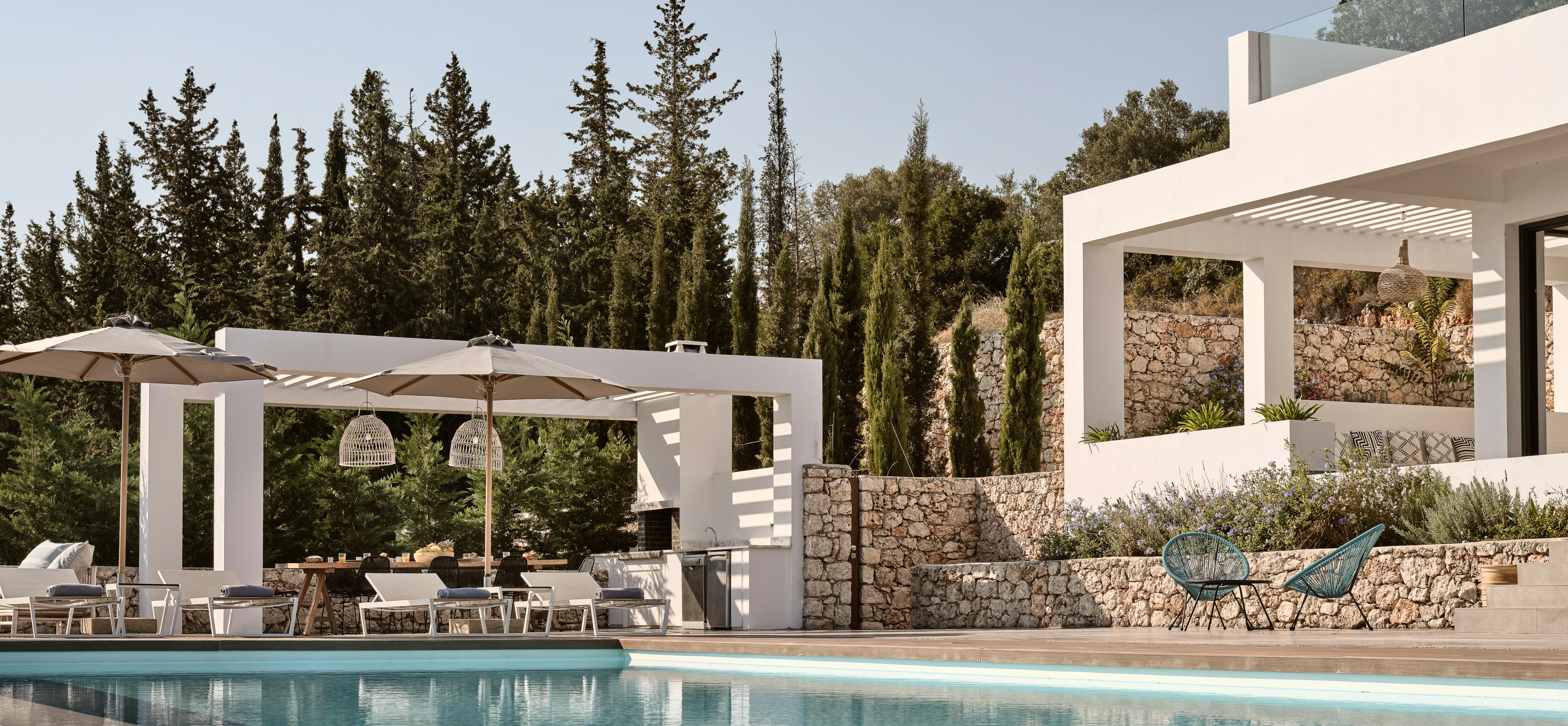 White minimalist villa exterior with infinity pool, pergola dining area, stone terracing, and Mediterranean cypress trees.