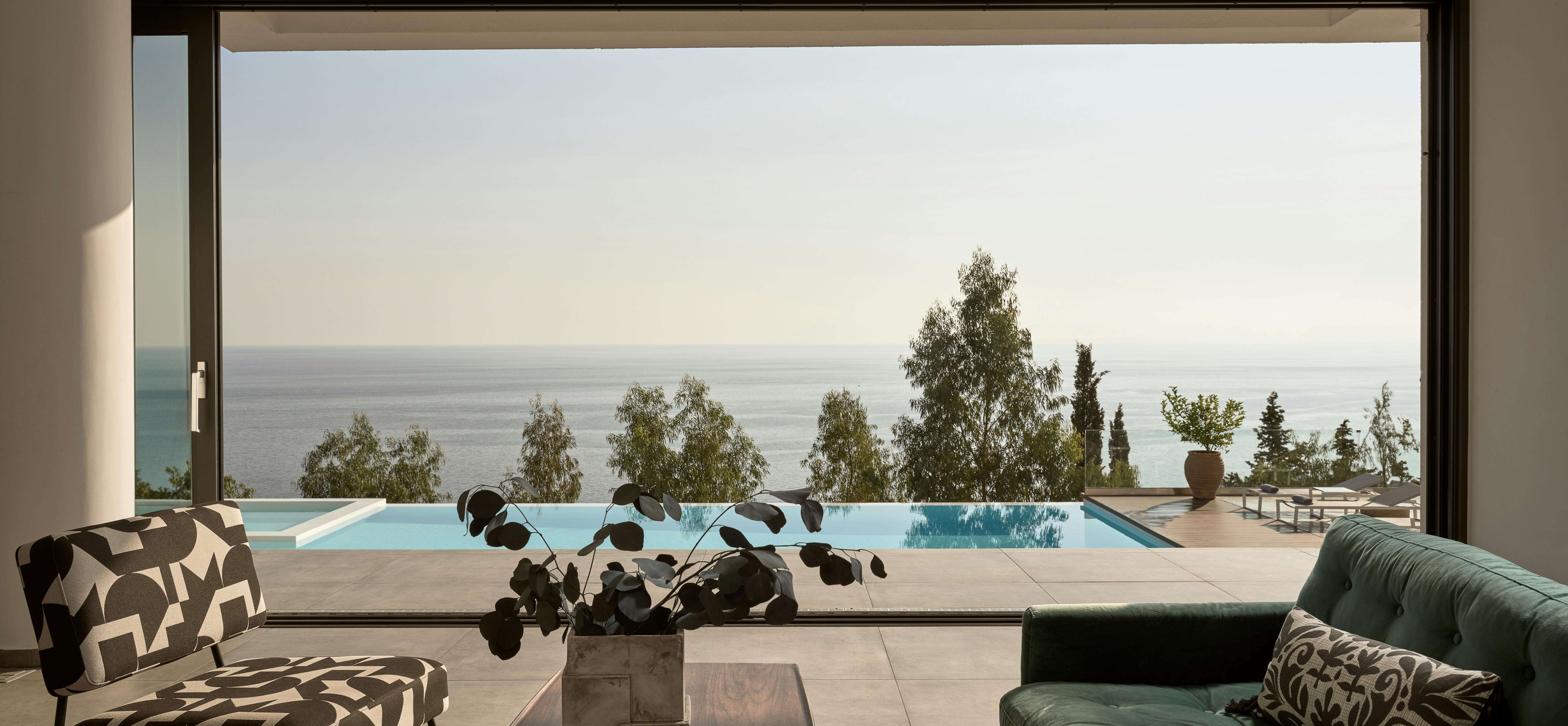 Modern living room with green velvet sofa, geometric chairs, and panoramic window framing infinity pool and sea view.