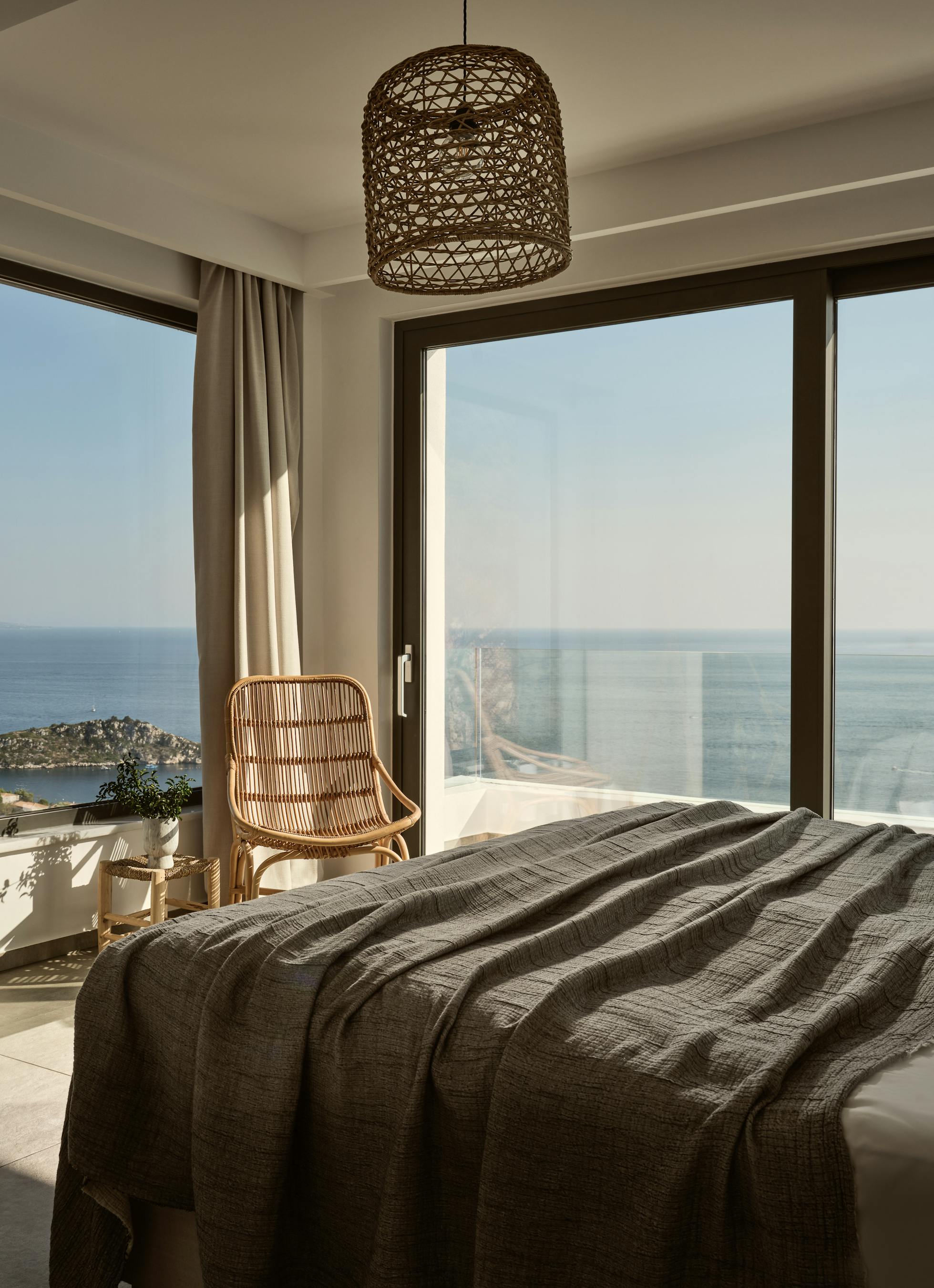 Bright bedroom with woven pendant light, rattan lounge chair, floor mirror, and corner windows framing coastal island views.