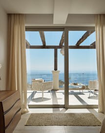 Contemporary bedroom with sculptural cream vase, wooden floating desk, and floor-to-ceiling glass doors opening to white wicker terrace furniture and ocean views.