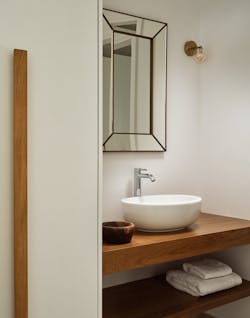 Modern bathroom with geometric brass-framed mirror, white vessel sink on natural wood floating shelf with lower storage, and neutral color palette.