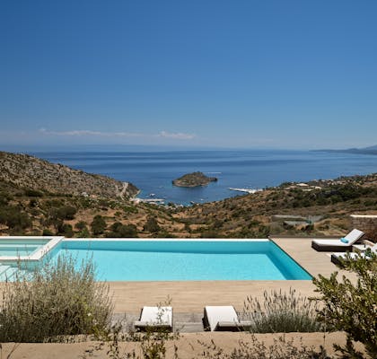 Spectacular infinity pool with wooden deck, white sun loungers, lavender plantings, and panoramic views of protected bay with island and anchored sailboats.