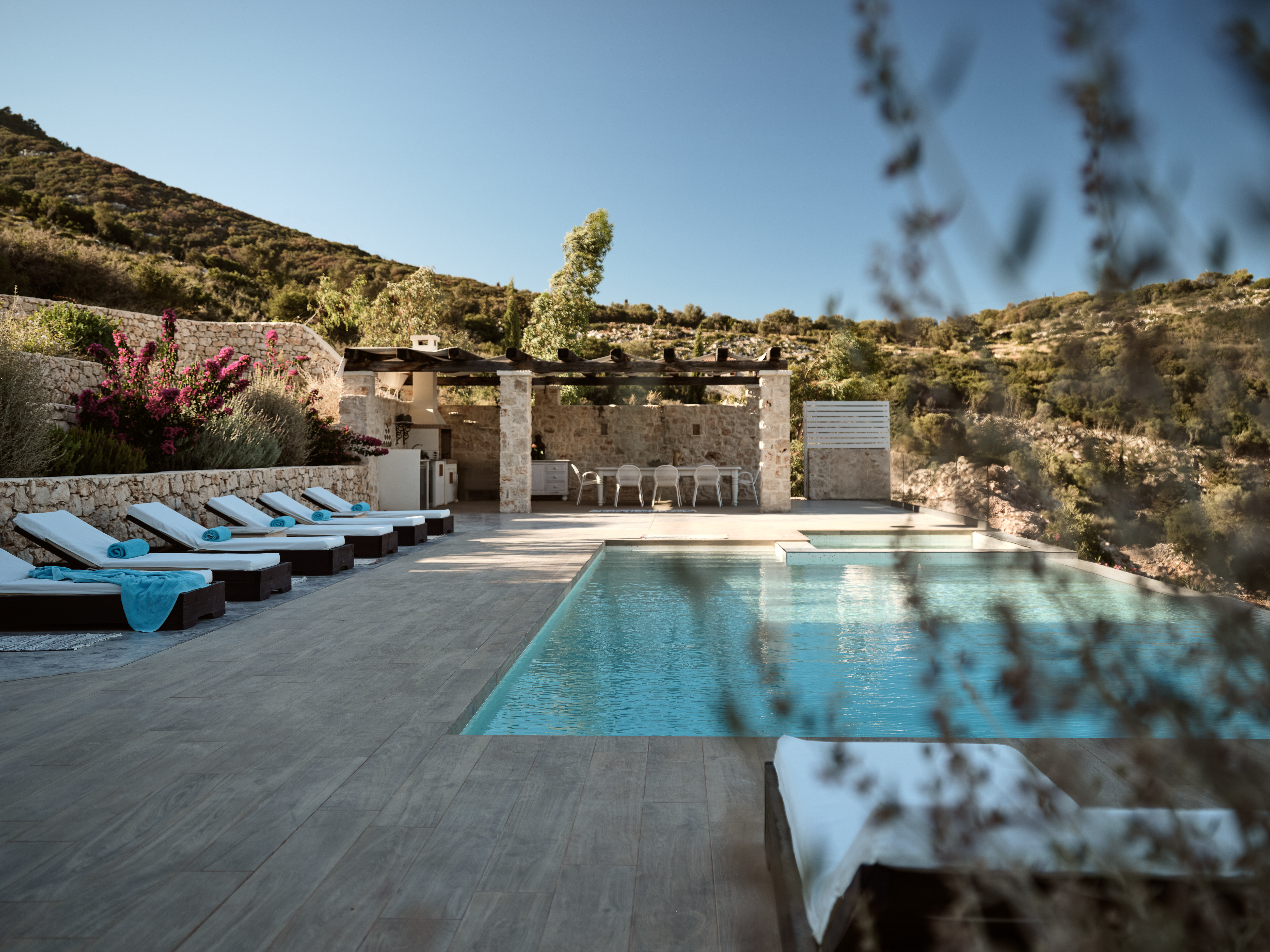 Hillside pool terrace with infinity edge, white sun loungers with turquoise towels, stone dining pavilion, and rugged Mediterranean landscape framed by vegetation.