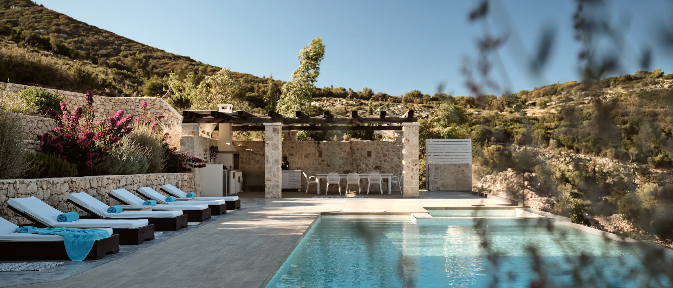 Hillside pool terrace with infinity edge, white sun loungers with turquoise towels, stone dining pavilion, and rugged Mediterranean landscape framed by vegetation.