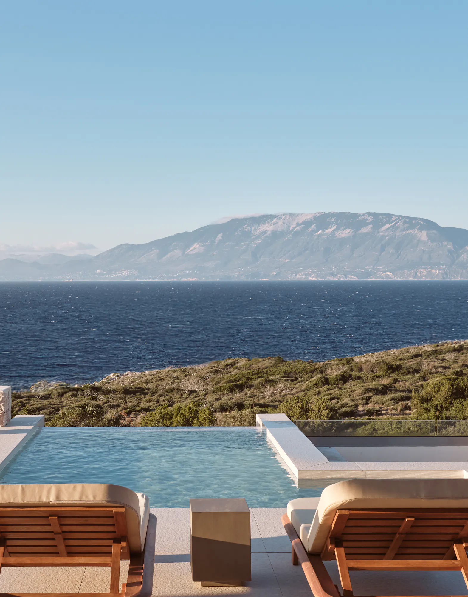 Covered terrace with wooden loungers overlooking an infinity pool and panoramic sea views toward distant mountains under clear blue sky.