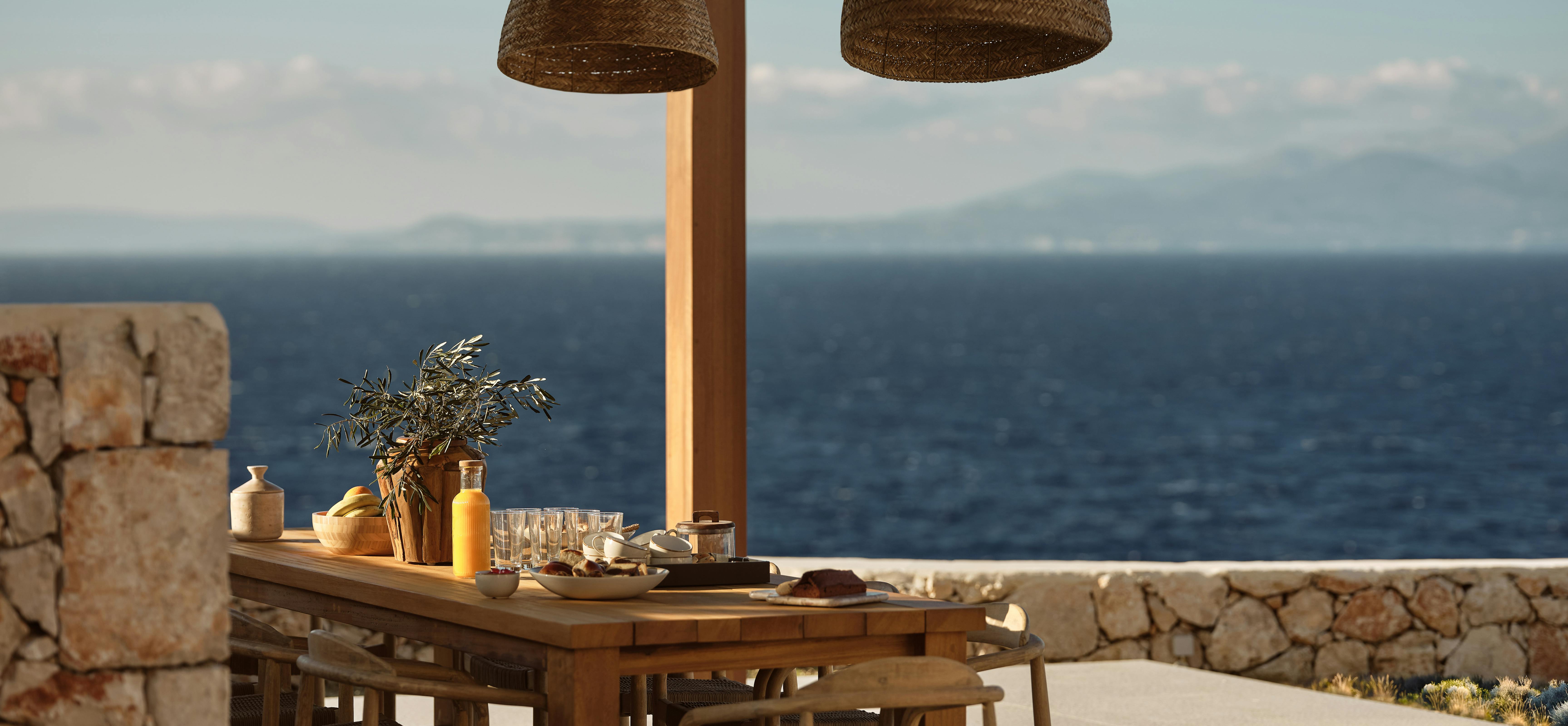 Outdoor dining area under wooden pergola with woven pendant lights, set table with breakfast items, and panoramic Mediterranean Sea views at golden hour.