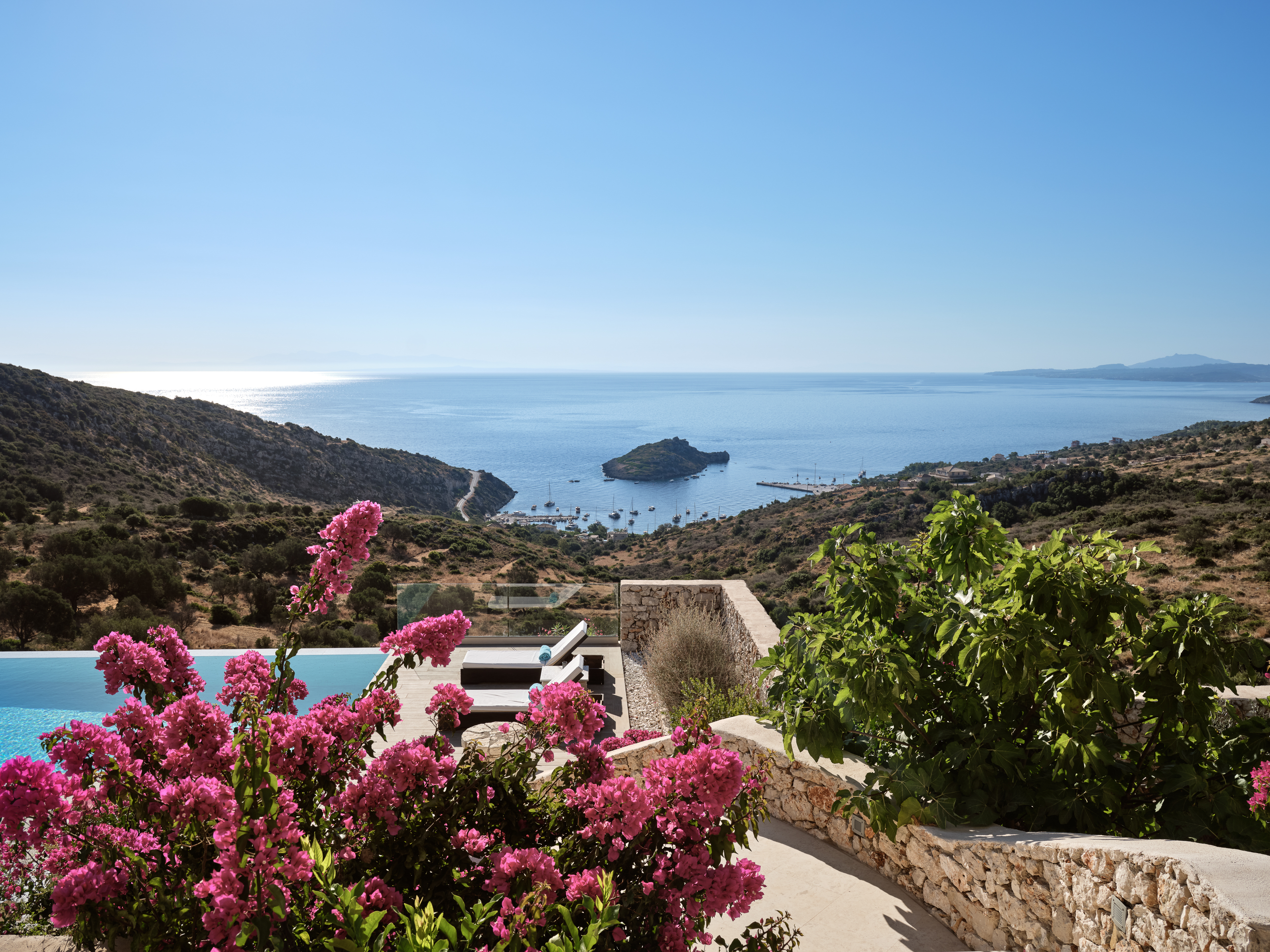 Dramatic hillside vista through bougainvillea with terraced infinity pool, modern villa architecture, protected bay with anchored boats, and distant islands.