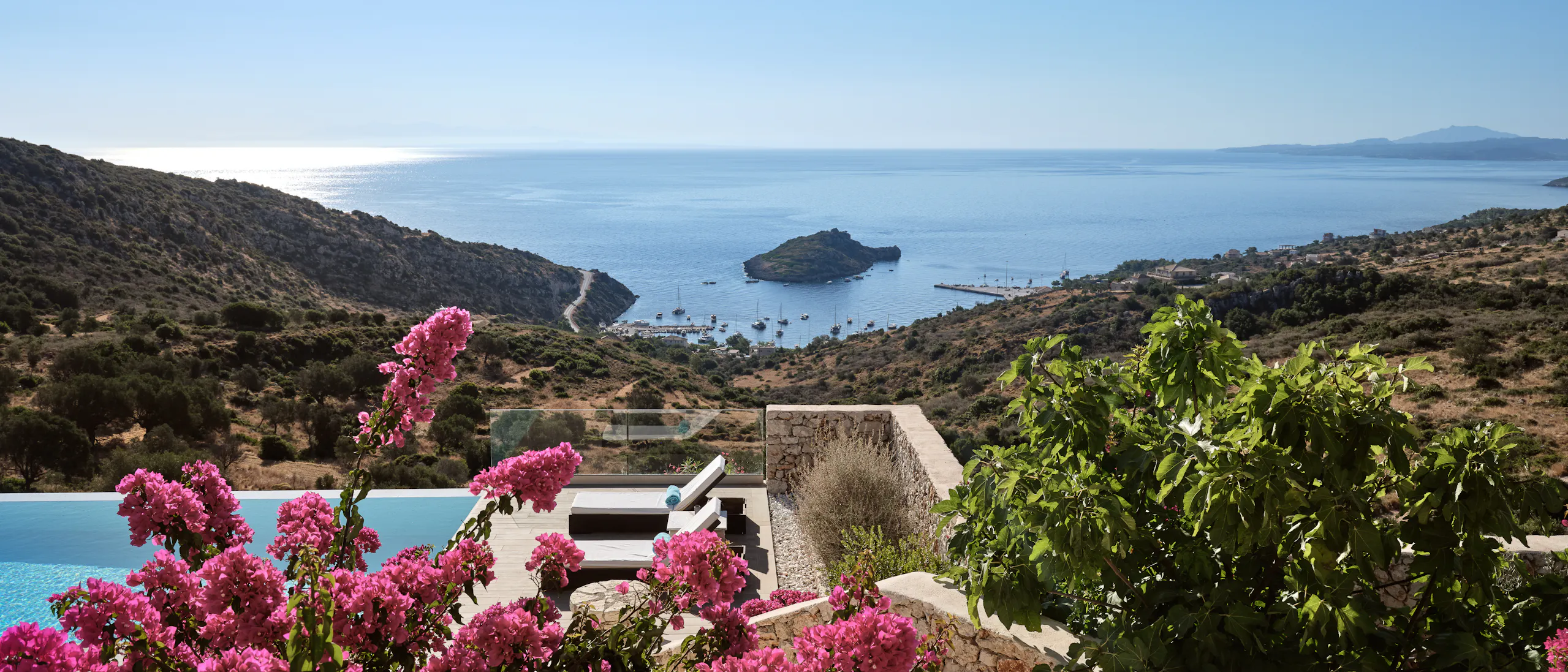 Dramatic hillside vista through bougainvillea with terraced infinity pool, modern villa architecture, protected bay with anchored boats, and distant islands.