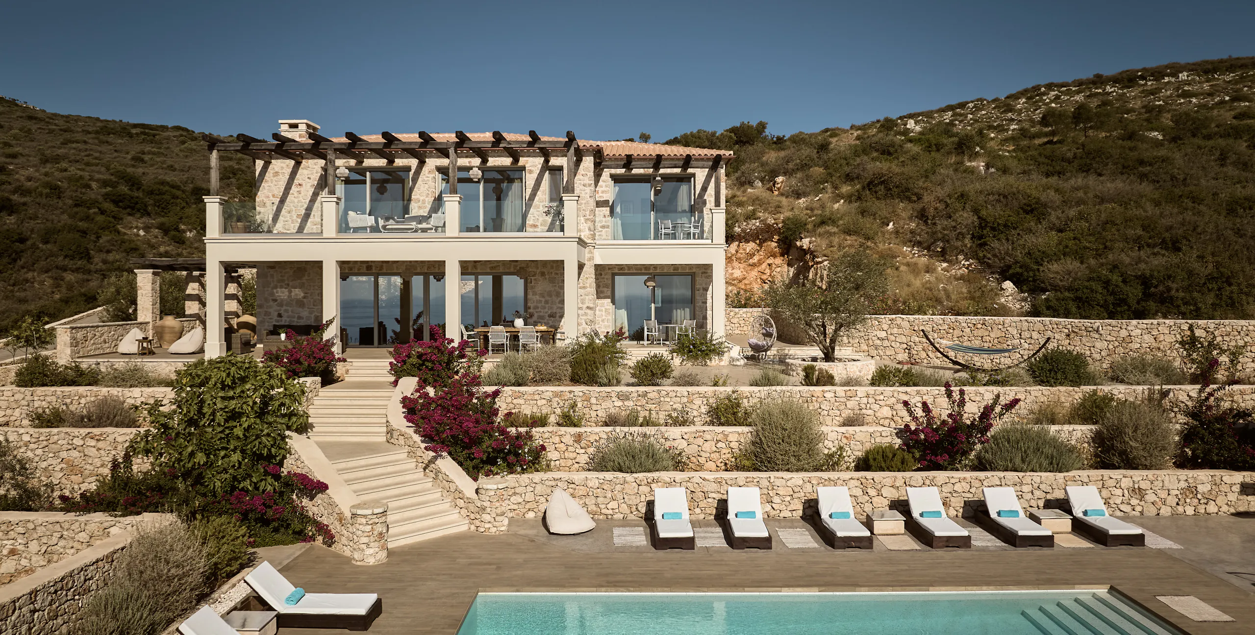 Two-story Mediterranean stone villa with wooden pergola rooftop terrace, terraced landscaping with bougainvillea, infinity pool, and sun loungers overlooking hillside.