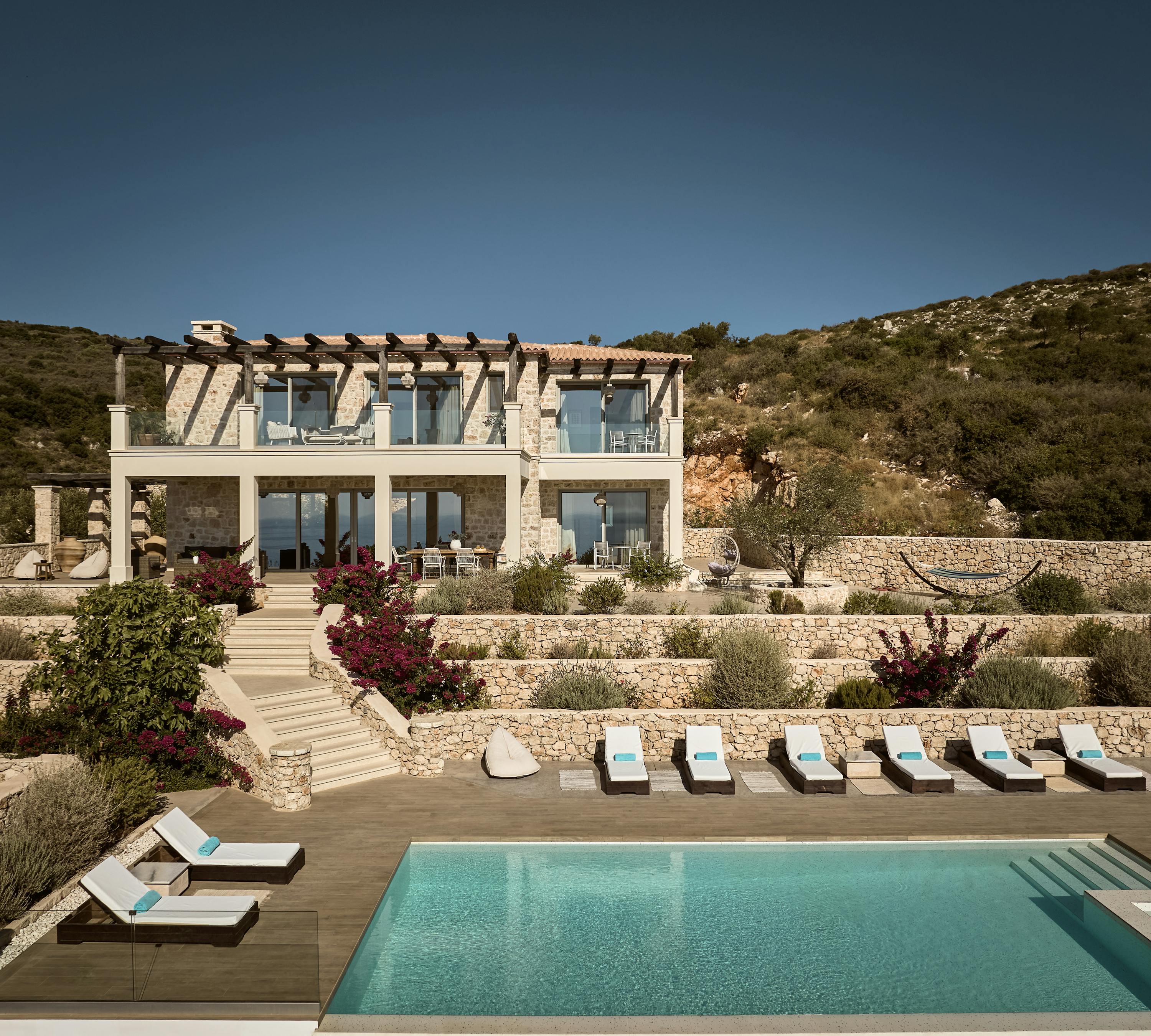 Two-story Mediterranean stone villa with wooden pergola rooftop terrace, terraced landscaping with bougainvillea, infinity pool, and sun loungers overlooking hillside.