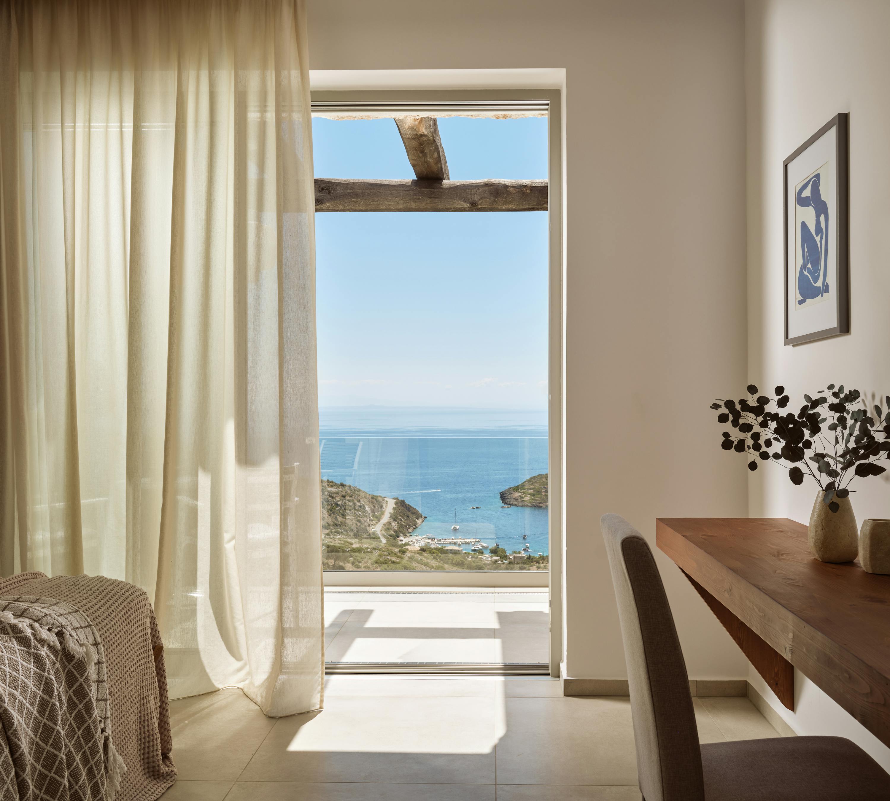 Sun-drenched bedroom featuring wooden furnishings, flowing curtains, and glass doors revealing wooden pergola terrace with bay and coastal hillside views.