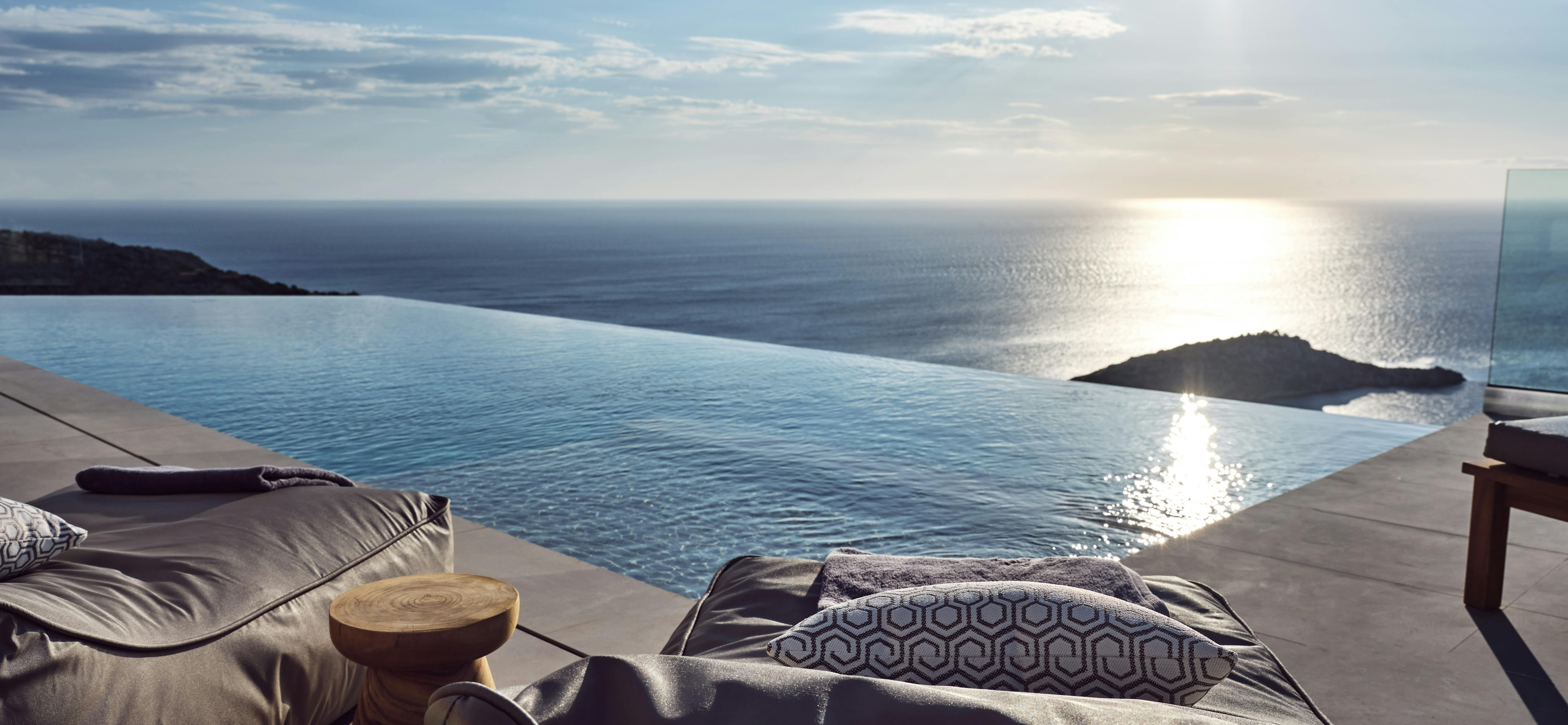 Infinity pool edge at sunset with dark loungers, geometric pillows, wooden side table, and dramatic sun reflection on water with silhouetted island.
