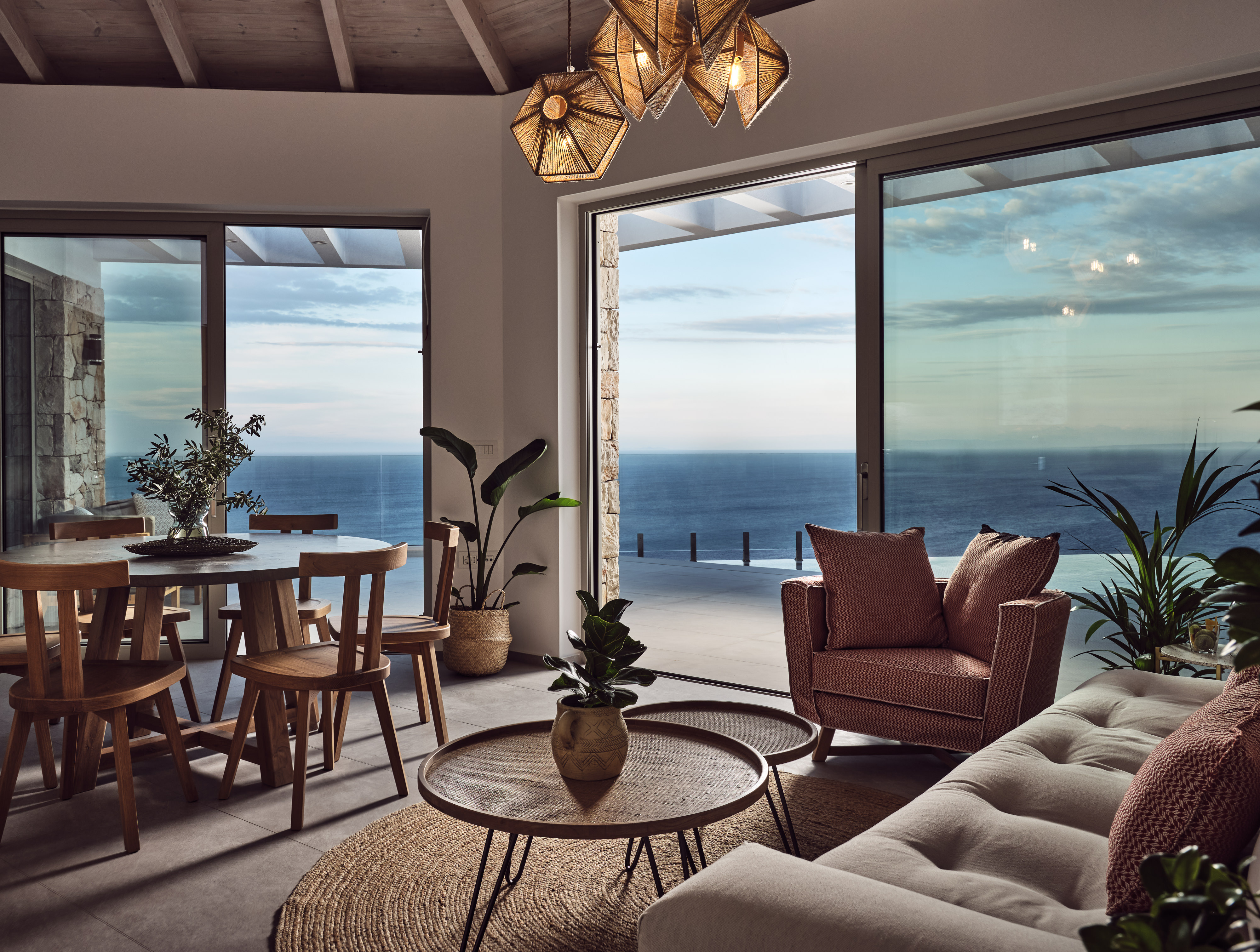 Open-plan living space with wooden dining set, terra-cotta textured armchair, tufted sofa, geometric pendant lights, and panoramic glass doors framing ocean and terrace views.