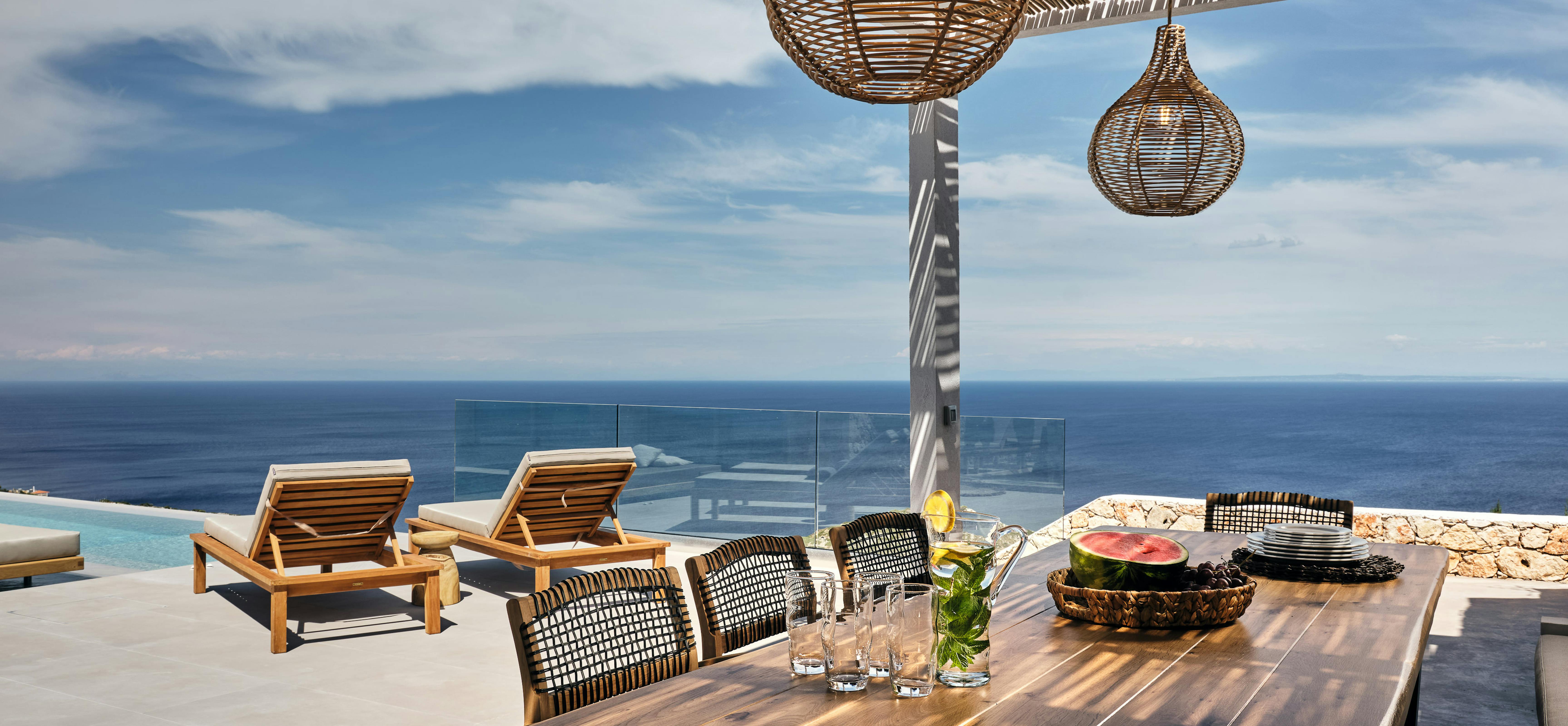 Outdoor dining terrace under slatted pergola with dark wooden table set with fresh watermelon, woven pendant lights, and infinity pool overlooking endless Mediterranean seascape.