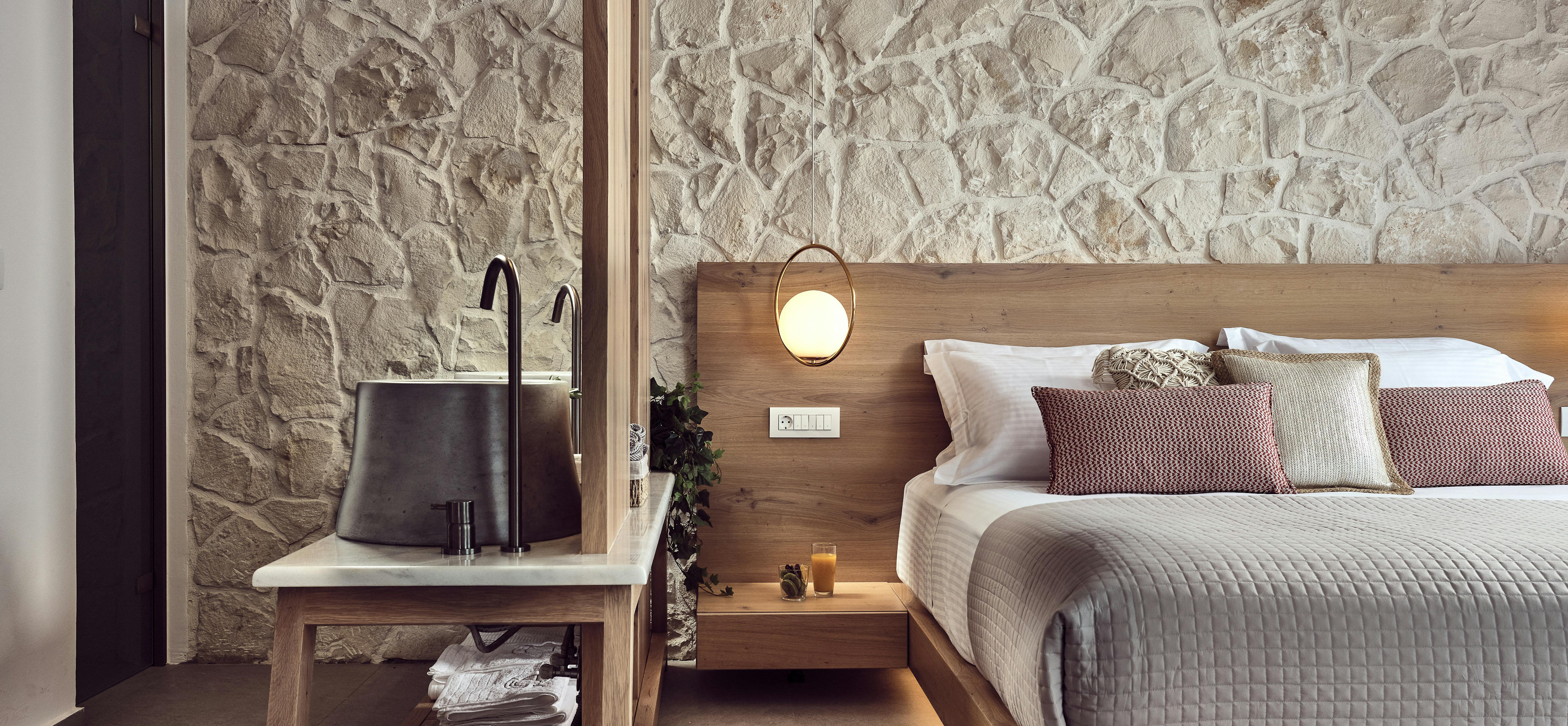 Modern bedroom detail with rough stone wall, wooden platform bed, white quilted bedding, floating nightstand with globe pendant light, and minimalist wooden vanity console.