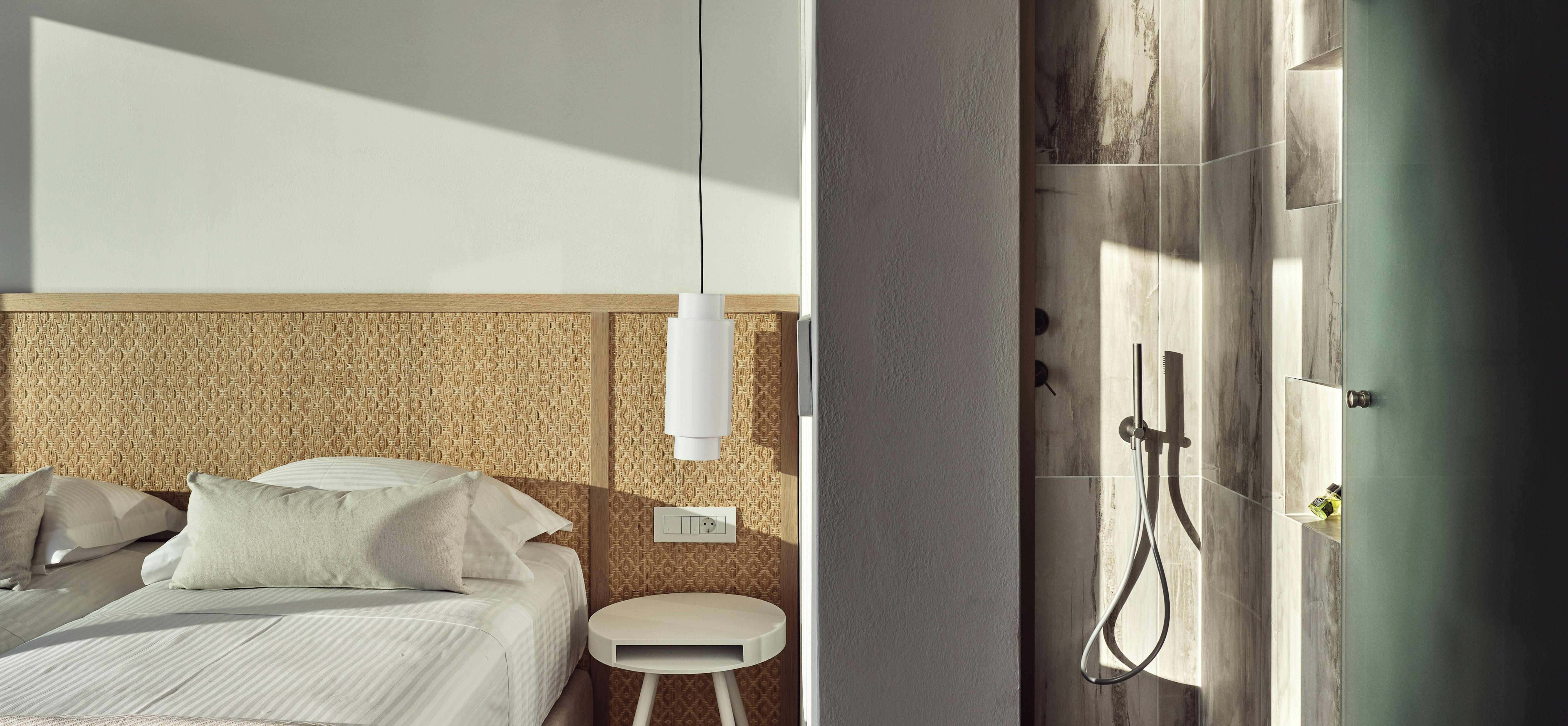 Minimalist bedroom with woven cane headboard wall panel, white cylindrical pendant light, round nightstand, and adjoining shower visible with wood-effect tiles and bronze fixtures.