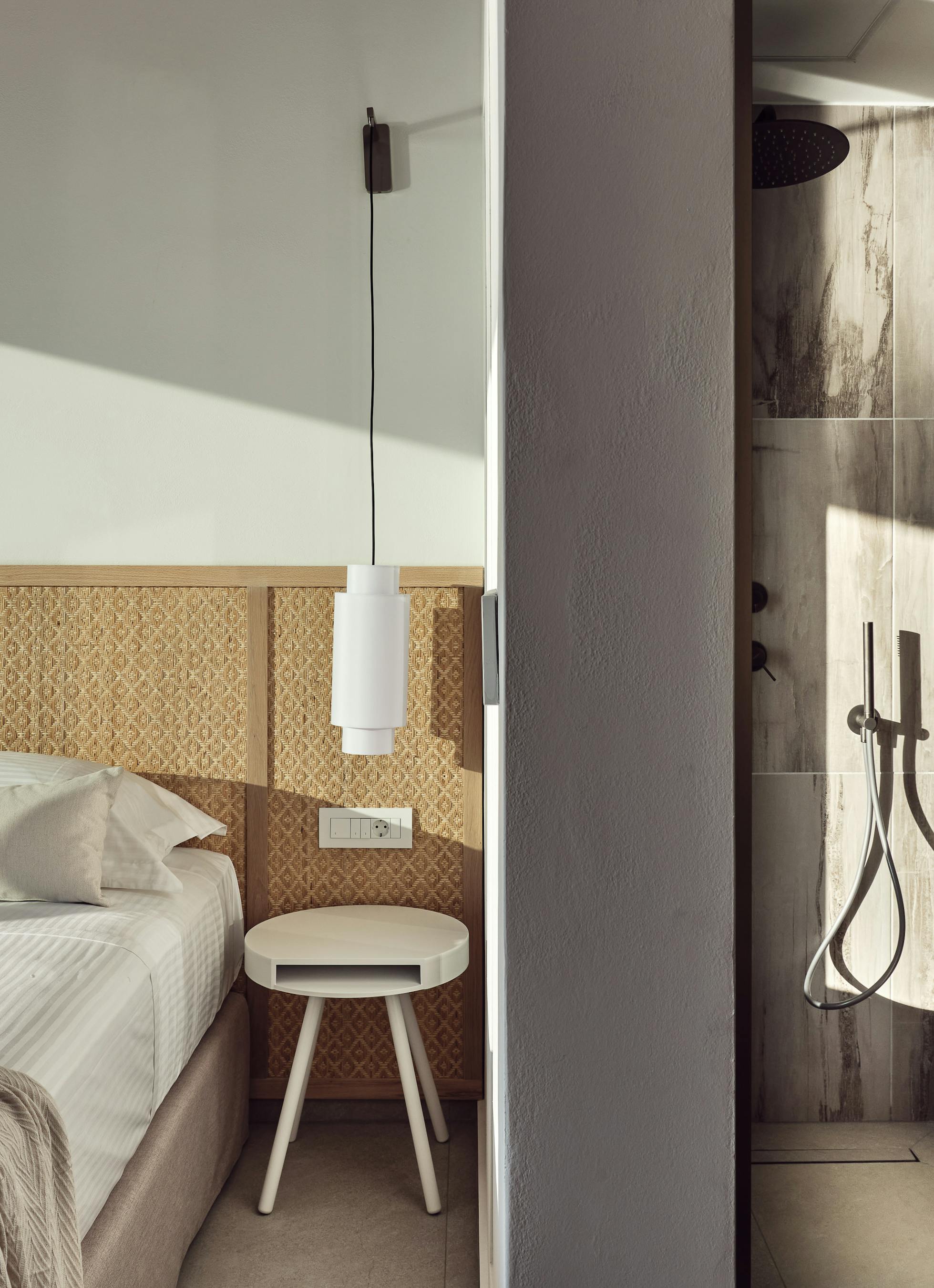 Minimalist bedroom with woven cane headboard wall panel, white cylindrical pendant light, round nightstand, and adjoining shower visible with wood-effect tiles and bronze fixtures.