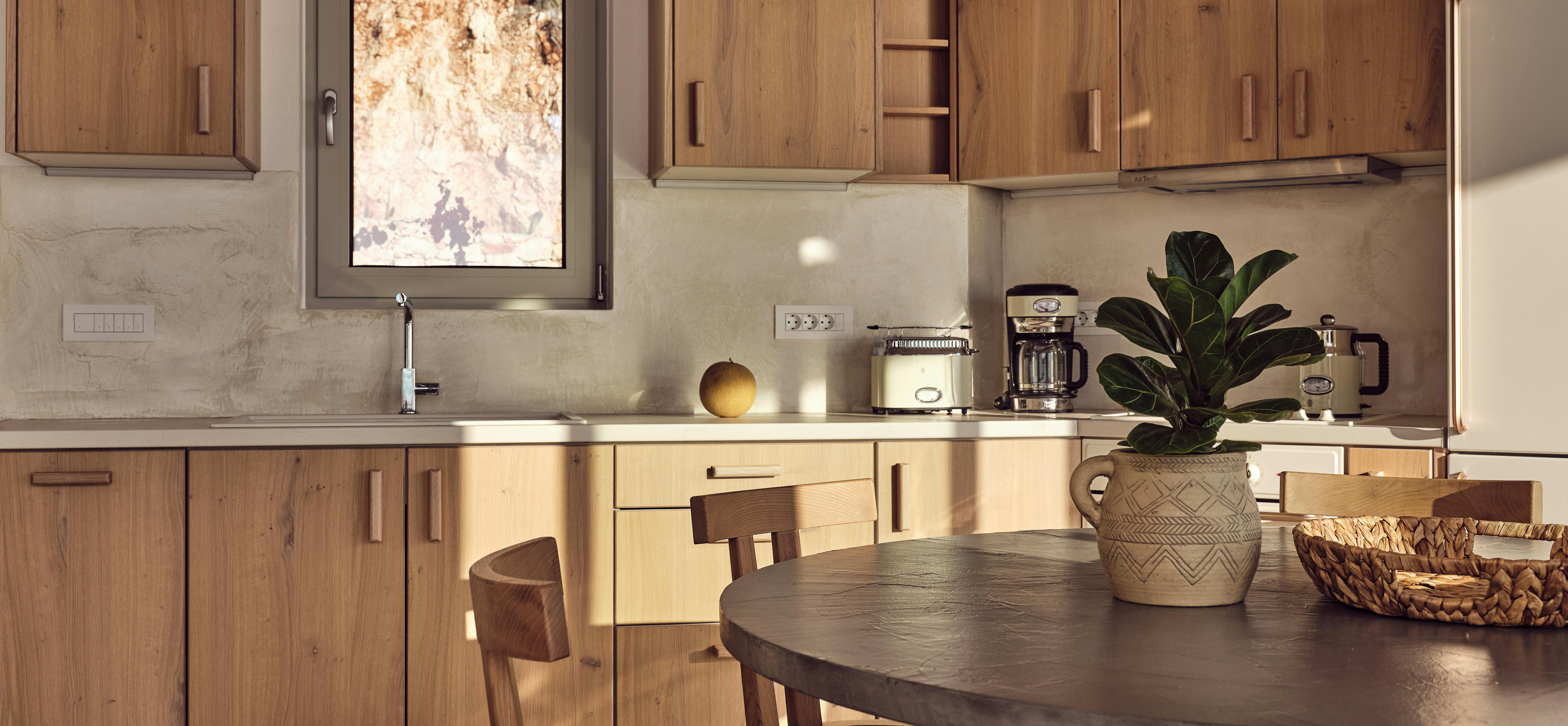 Compact kitchen bathed in warm sunlight with oak upper and lower cabinets, window framing rocky hillside, round dark wood dining table, and breakfast items.