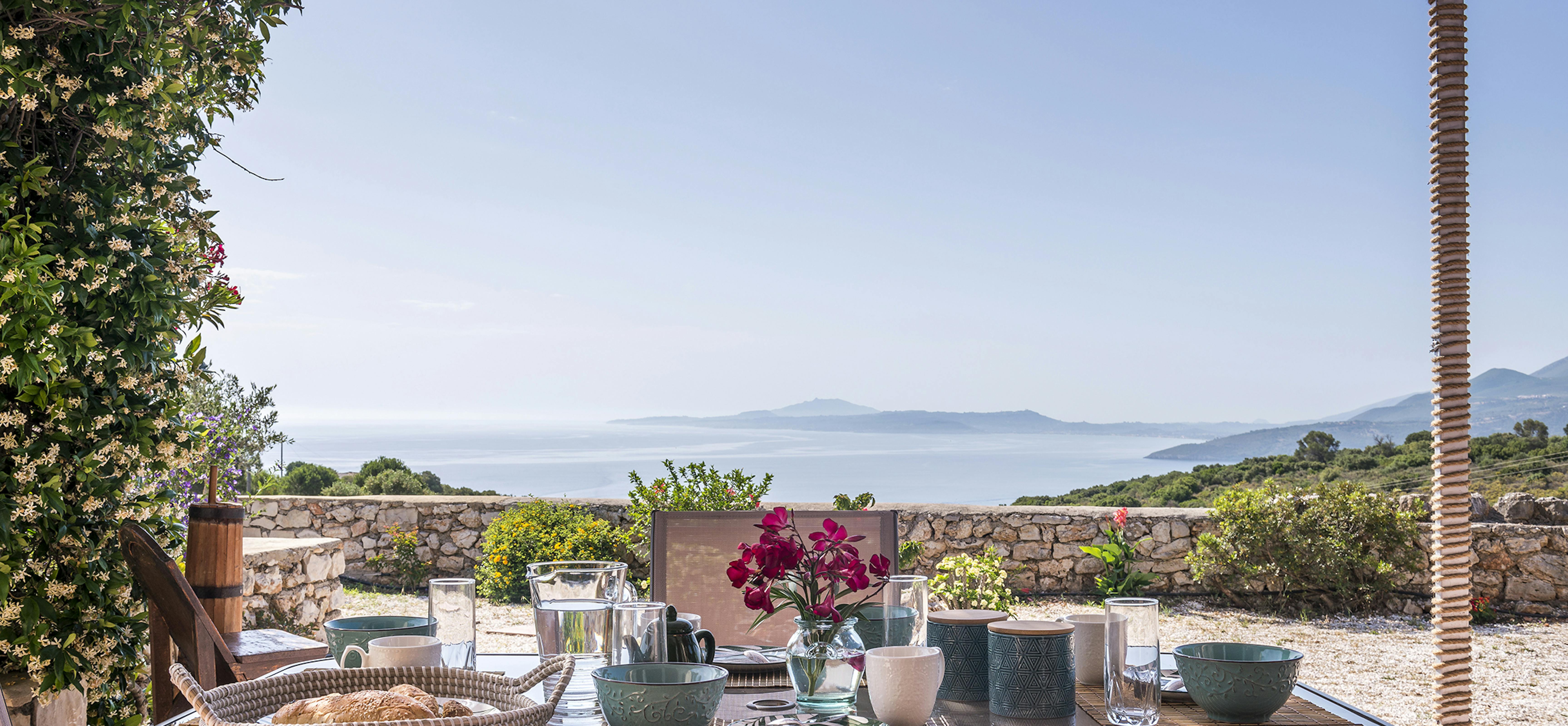 Outdoor breakfast table under reed pergola set with colourful ceramic bowls, glass water pitcher, fresh bougainvillea, and panoramic views of blue Mediterranean Sea and distant islands.