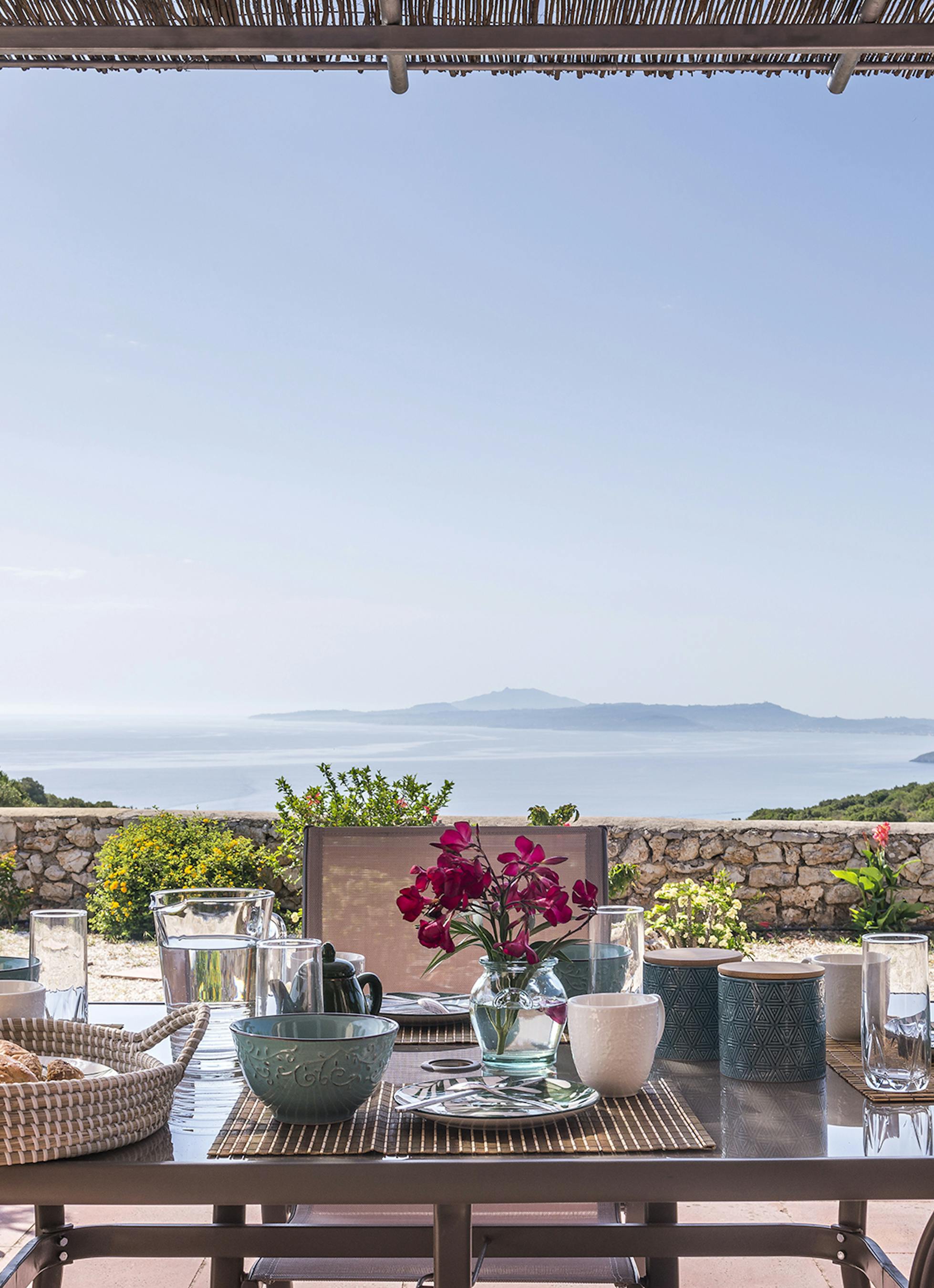 Outdoor breakfast table under reed pergola set with colourful ceramic bowls, glass water pitcher, fresh bougainvillea, and panoramic views of blue Mediterranean Sea and distant islands.