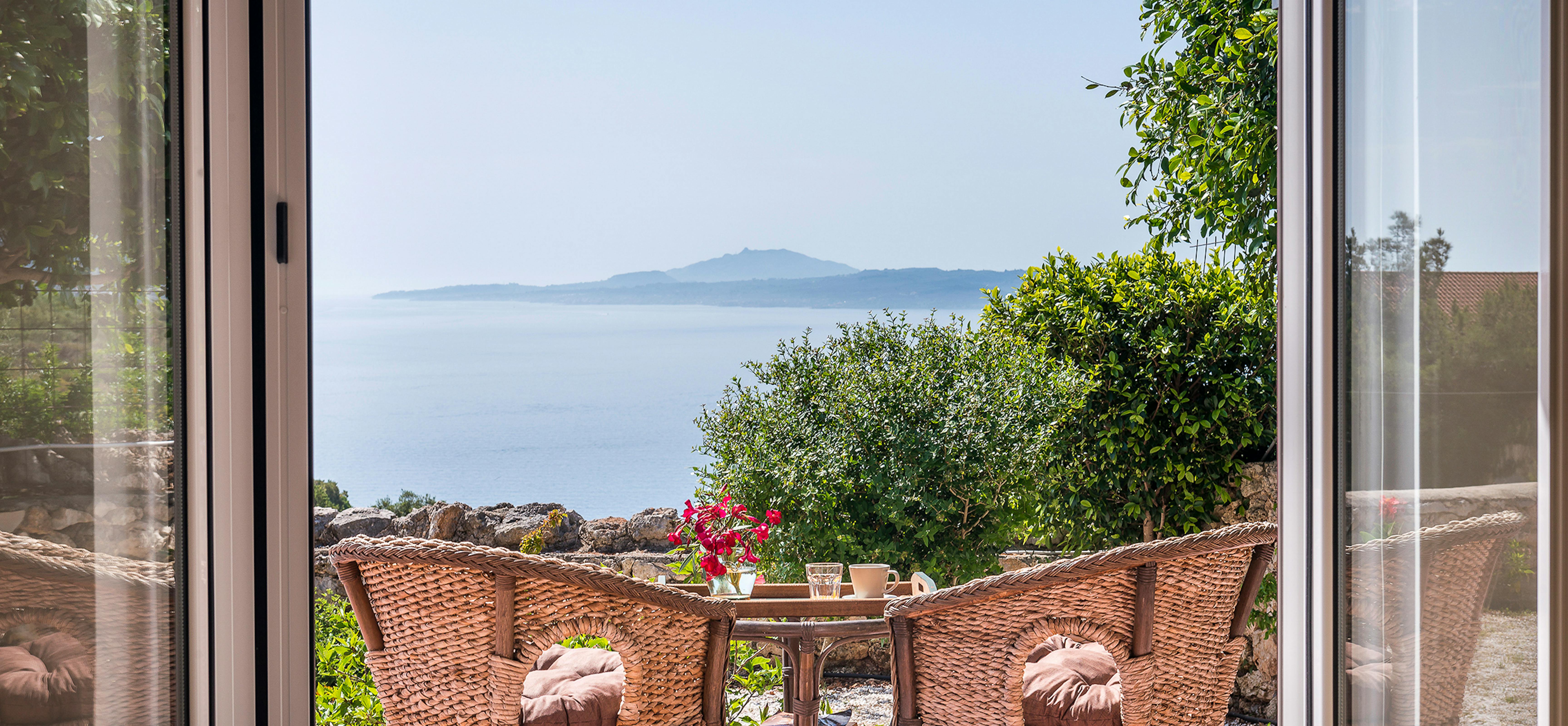 Private breakfast terrace framed by sliding doors with wicker chairs, rustic wooden table set with coffee and bougainvillea, overlooking dramatic coastal cliffs and azure sea.