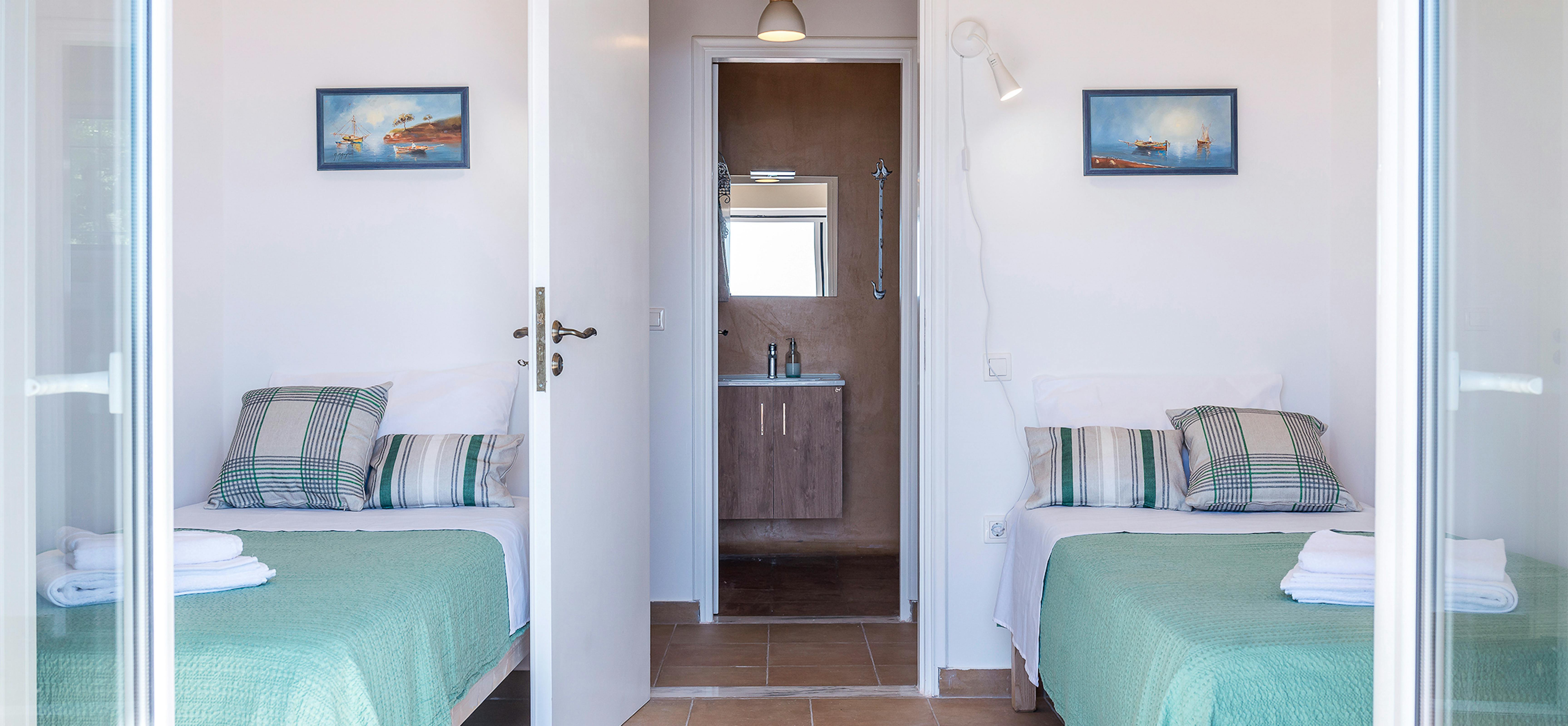 Bright twin bedroom with mirrored walls, aqua quilted bedspreads, striped nautical pillows, matching boat paintings, and view through to compact bathroom with dark wood vanity.