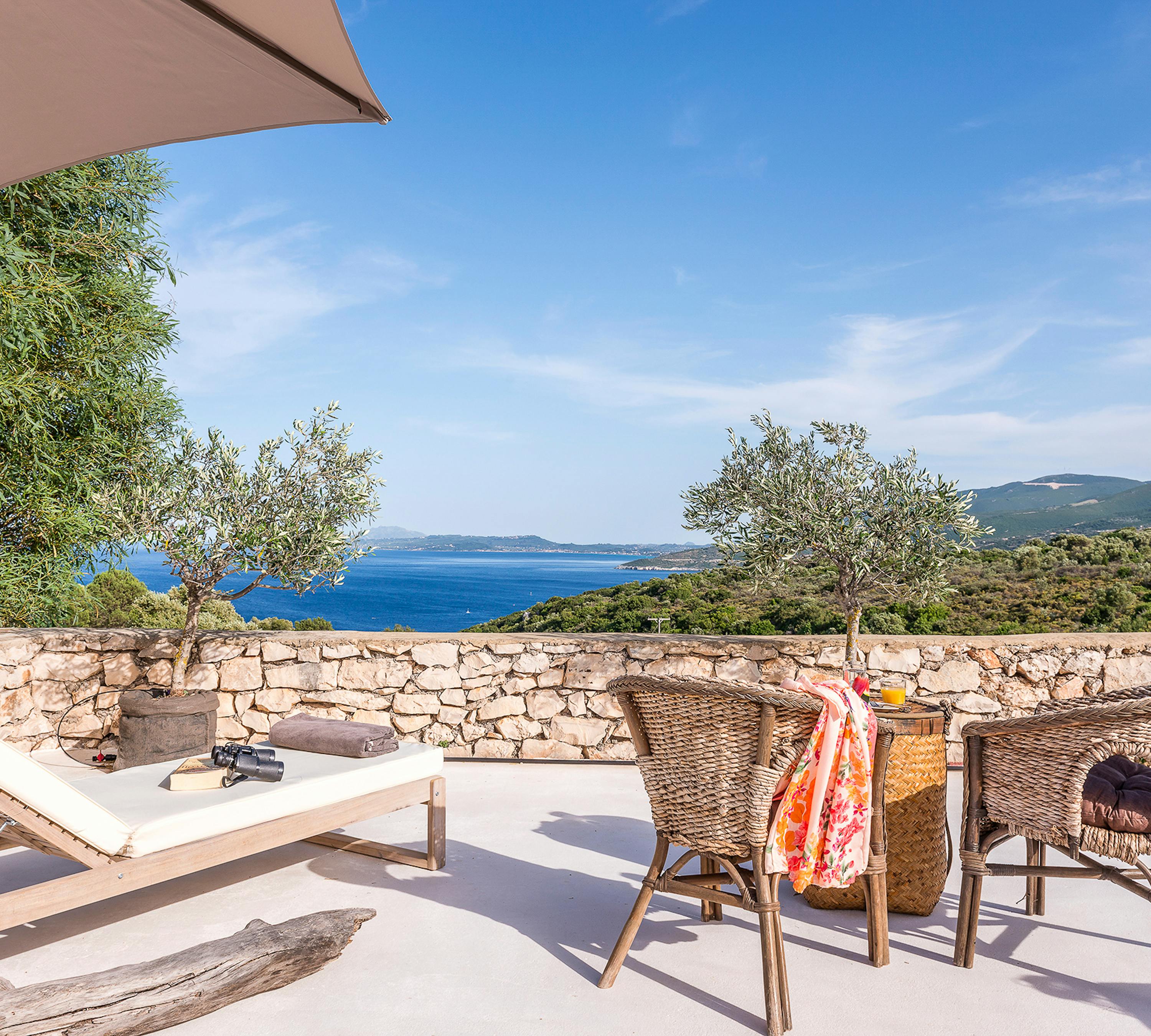 Sun-drenched terrace with white lounger, wicker dining set draped with colourful towel, taupe umbrella, stone retaining walls, olive trees, and sweeping coastal panorama.