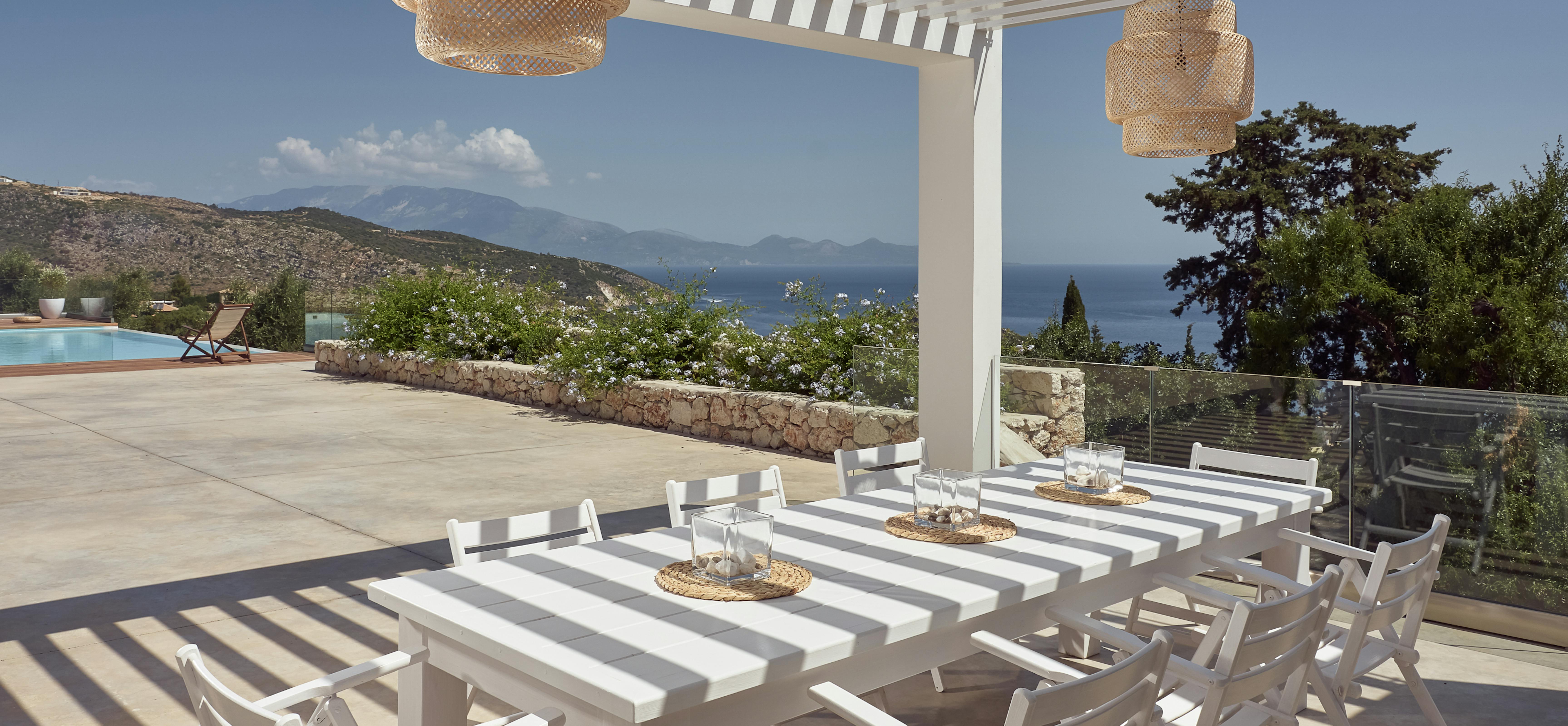 Outdoor dining terrace with long white table under striped pergola overlooking mountains and sea.
