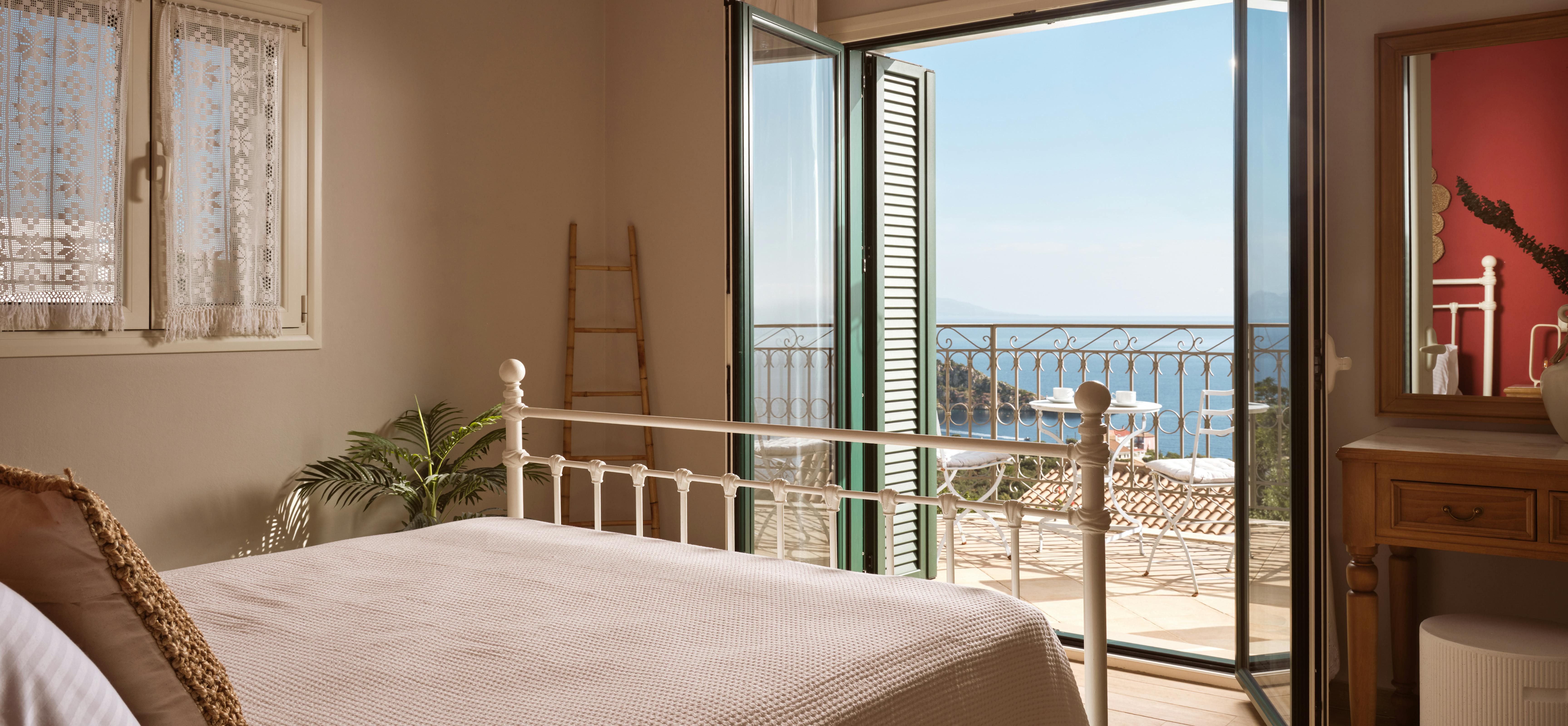 A bedroom with a white iron bed frame opens to a sea-view balcony through French doors. Traditional lace curtains and wooden furniture complement the coastal Mediterranean setting.