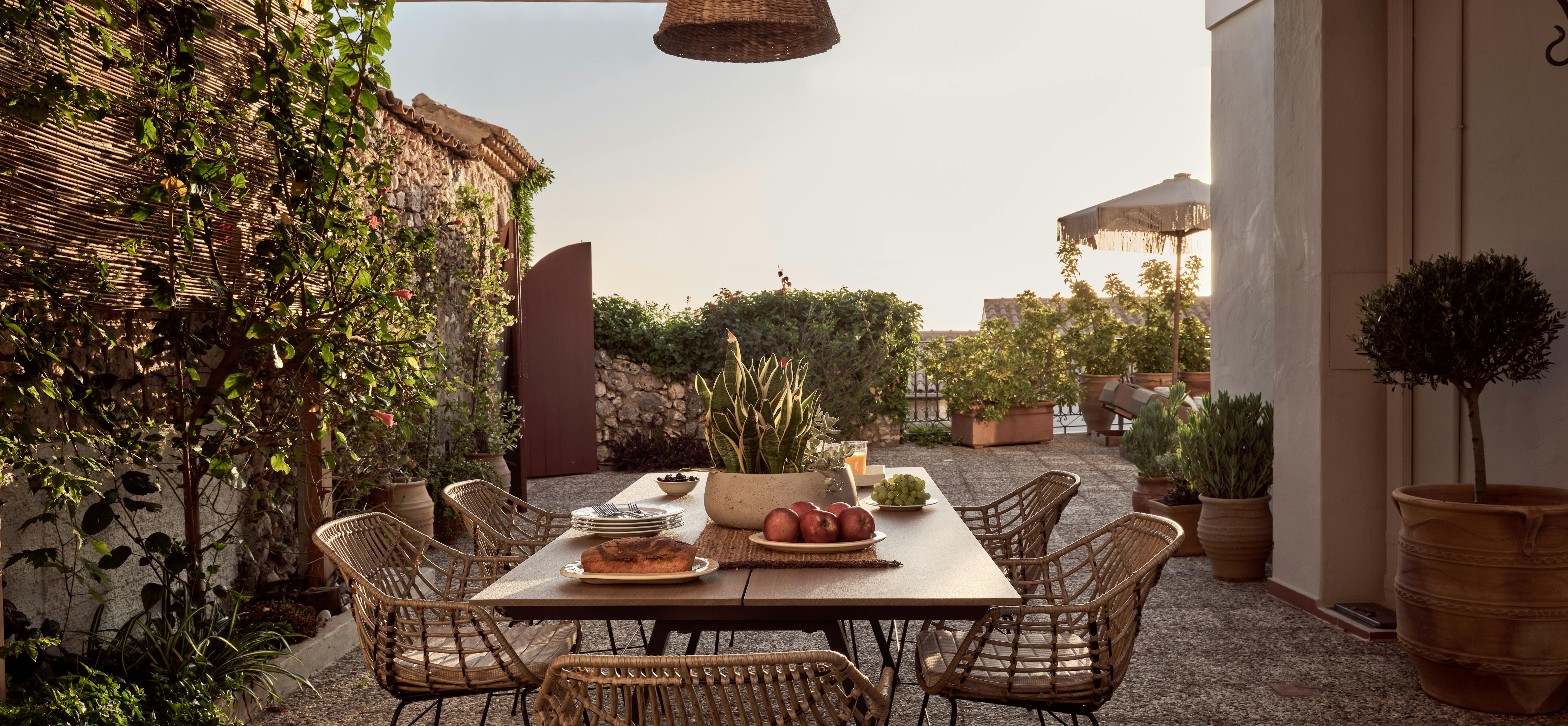 A covered outdoor dining area features rattan chairs around a wooden table under a pergola. Terracotta pots and climbing vines create an intimate Mediterranean courtyard atmosphere.