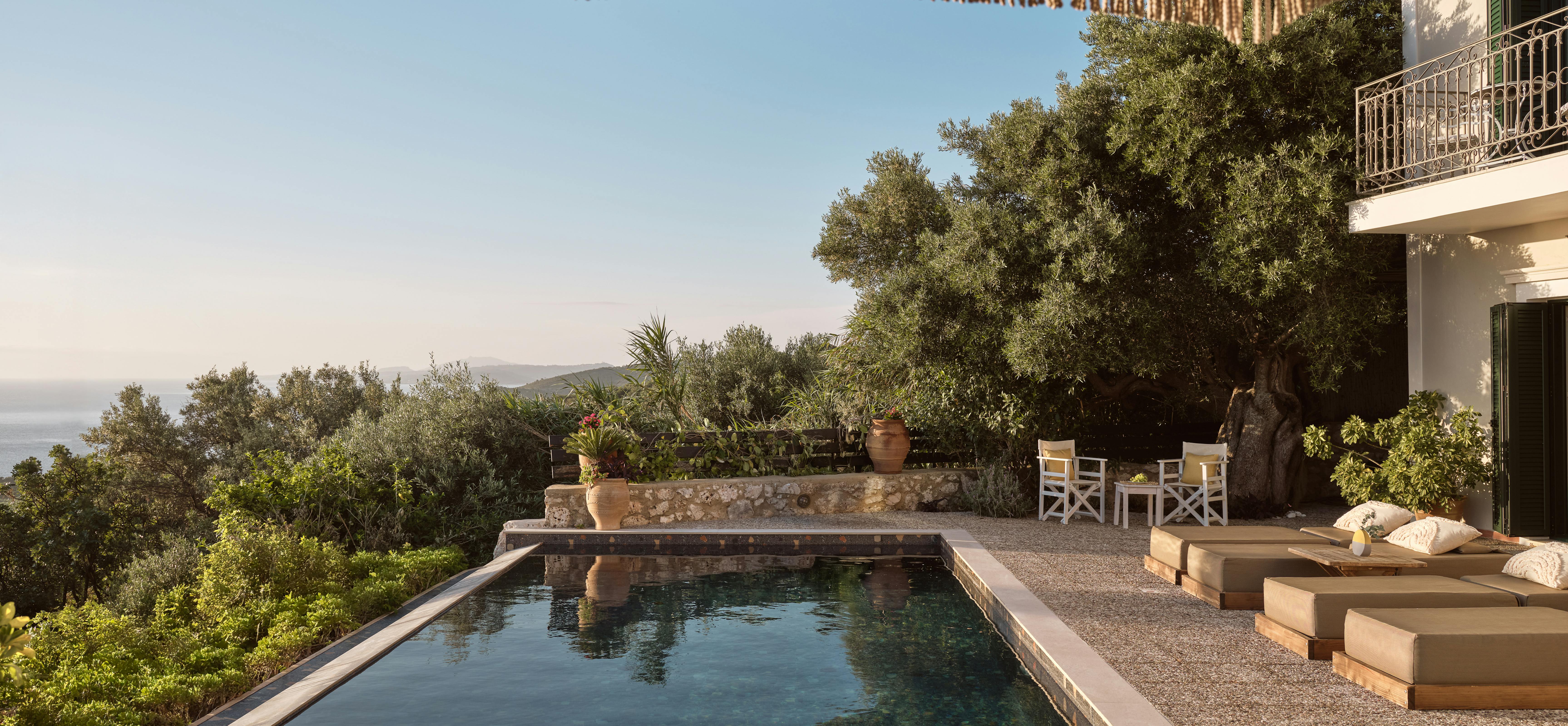 A hillside swimming pool terrace overlooks a panoramic landscape with mountains and sea views. Modern lounge furniture and a fringed umbrella create an elegant outdoor living space surrounded by Mediterranean vegetation.