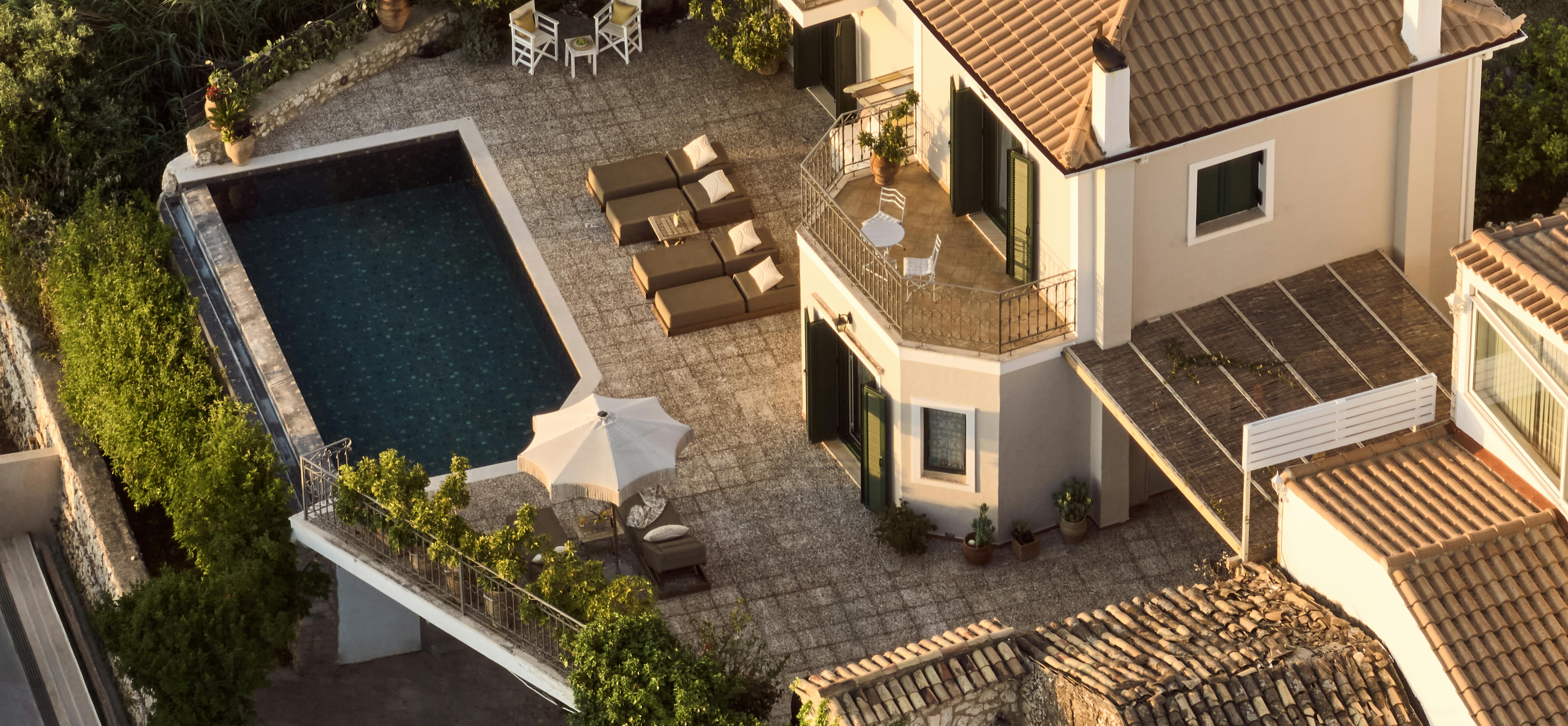 An aerial view shows a Mediterranean villa with terracotta roofing surrounded by a rectangular swimming pool. The terrace and balconies create outdoor living spaces among lush vegetation.