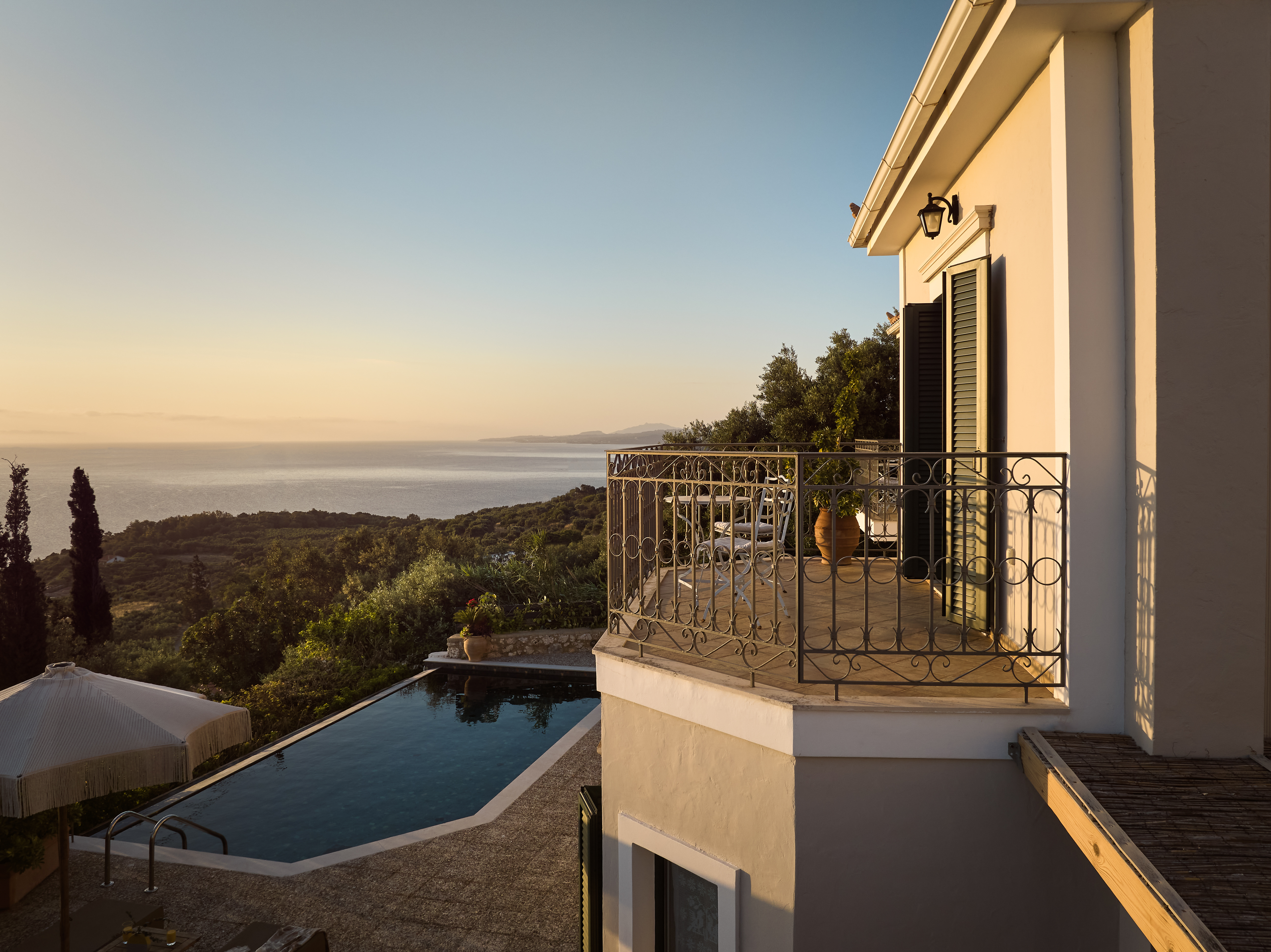 A villa balcony captures golden hour light with wrought iron railings overlooking rolling hills and distant sea. The terracotta-roofed building exemplifies classic Mediterranean architecture.