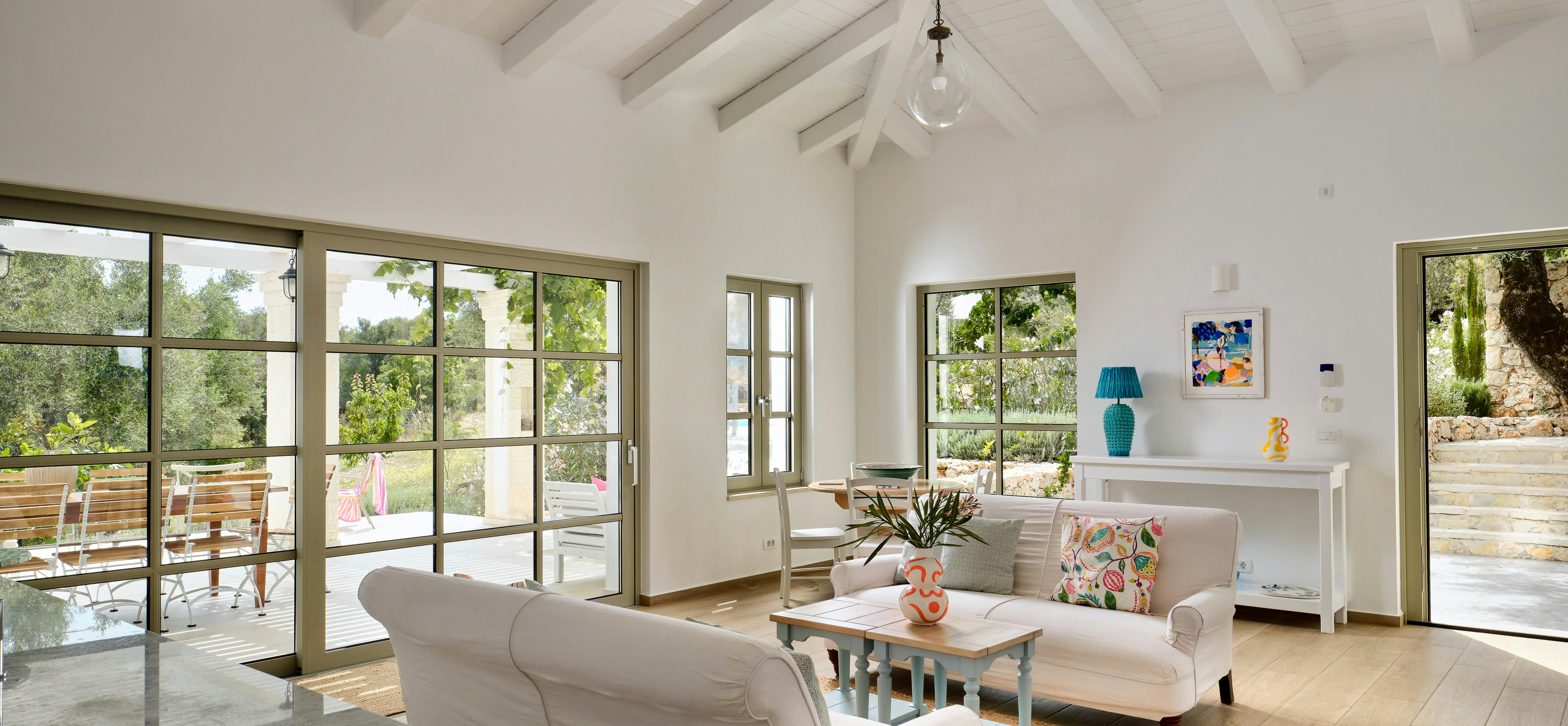 Open-plan living area with white sofas, exposed ceiling beams, and large windows. The space flows to outdoor terrace dining visible through glass doors.