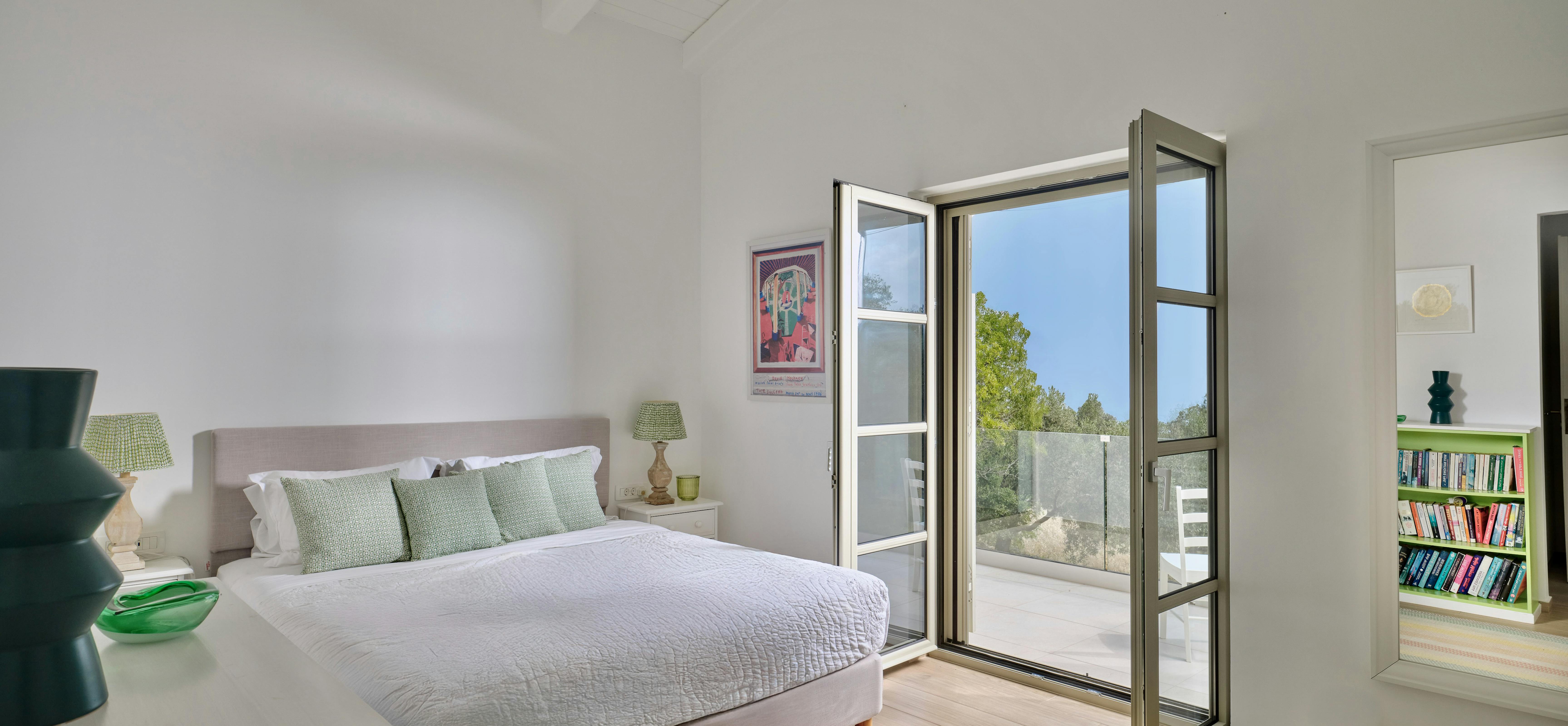 Master bedroom with neutral bedding and green accent pillows opening to private terrace. White ceiling beams and natural light create a serene atmosphere.