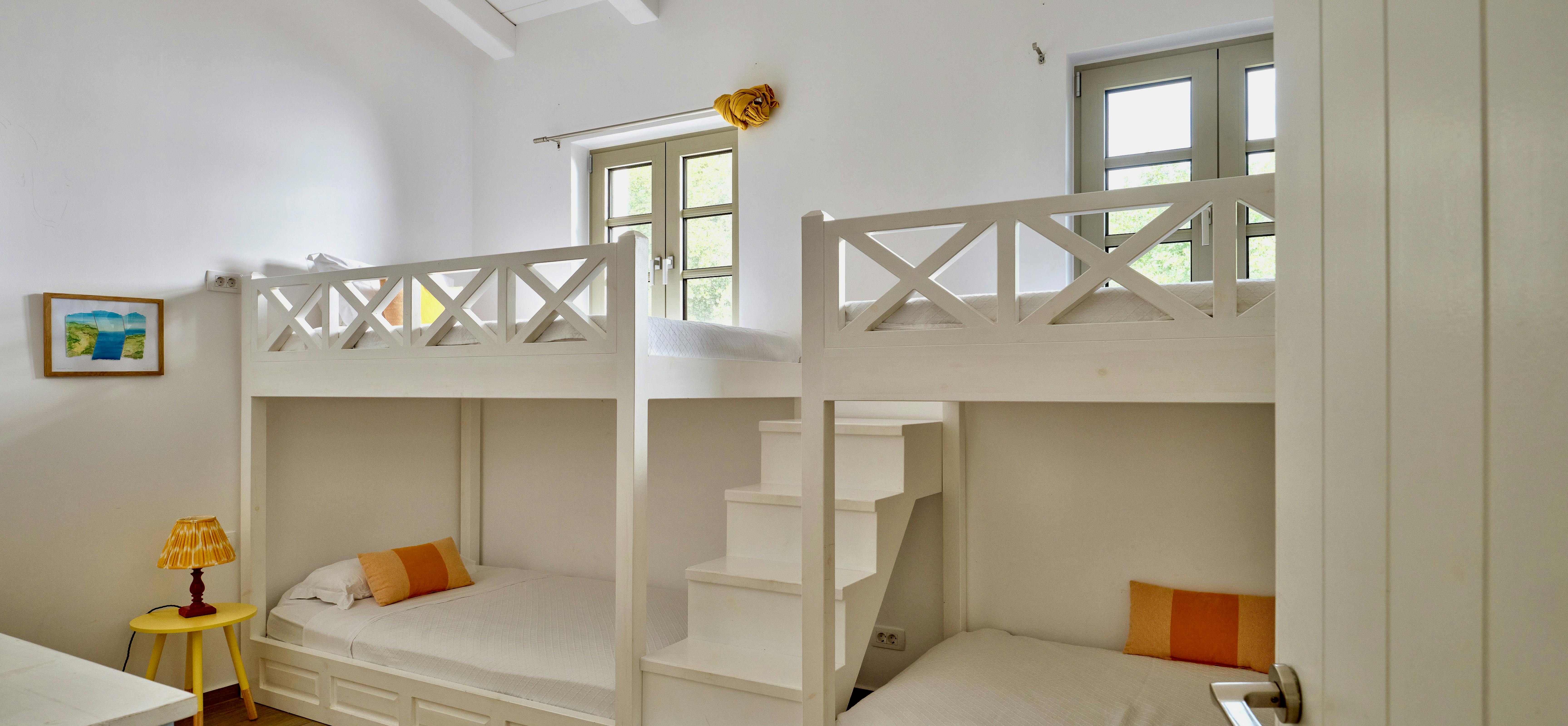 Children's bedroom with white built-in bunk beds featuring crossed railings and storage drawers. Exposed white ceiling beams and yellow accent pieces add warmth.