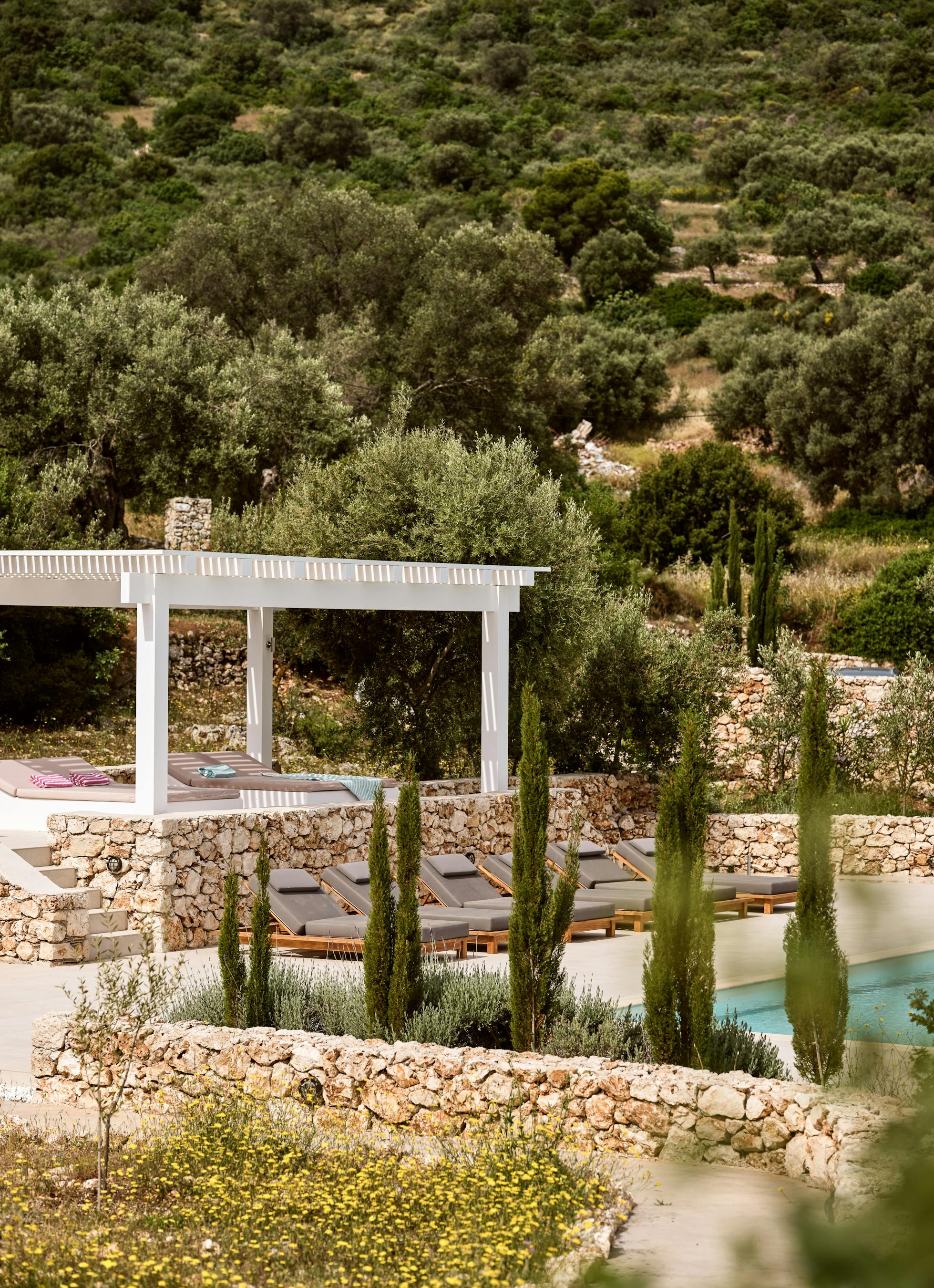 Pool area from elevated perspective showing integration with natural hillside terrain. Stone retaining walls and Mediterranean plantings blend architecture with landscape.