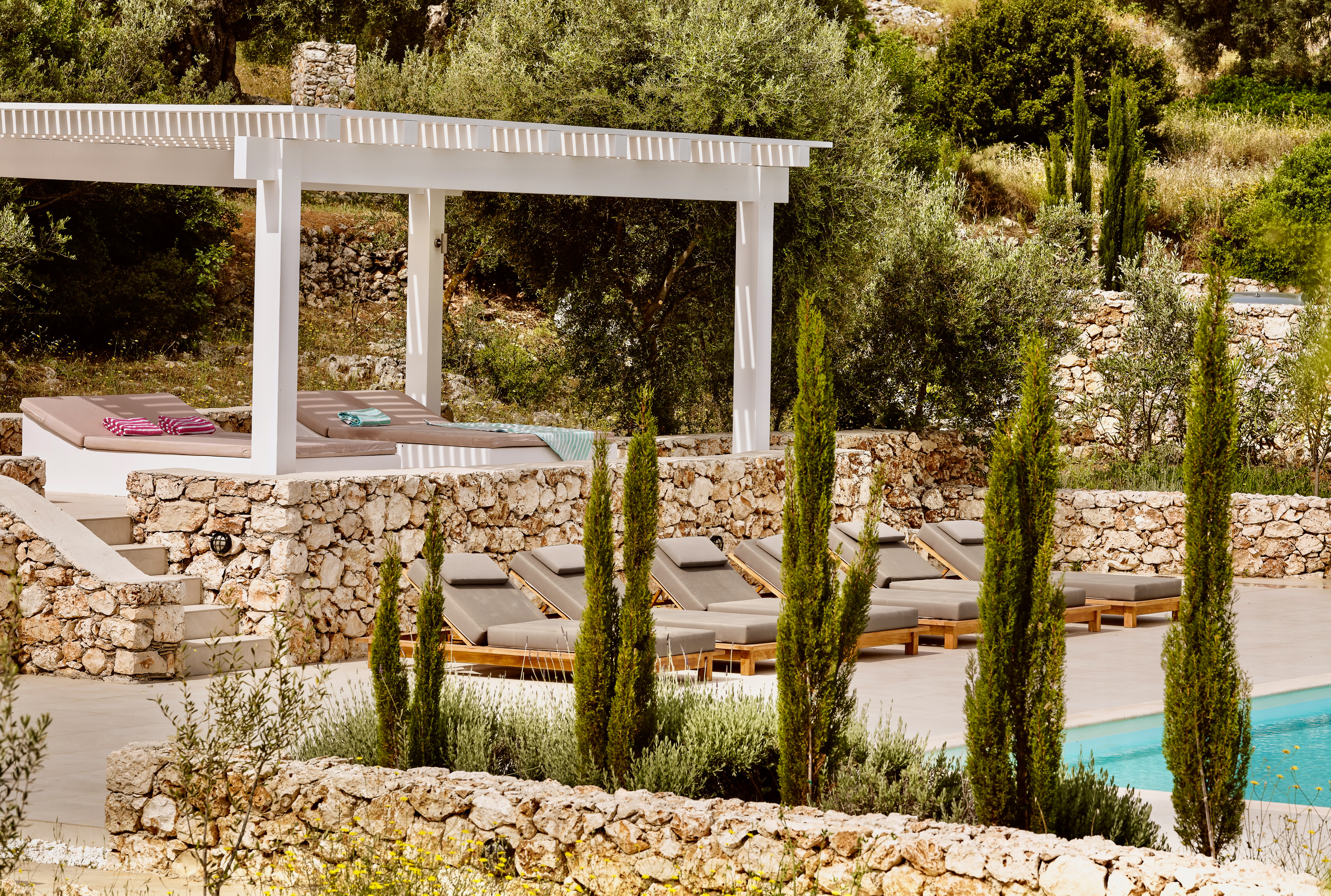 Multi-level pool deck with white pergola and wooden sun loungers with grey cushions. Tall cypress trees and stone walls define the terraced landscape design.