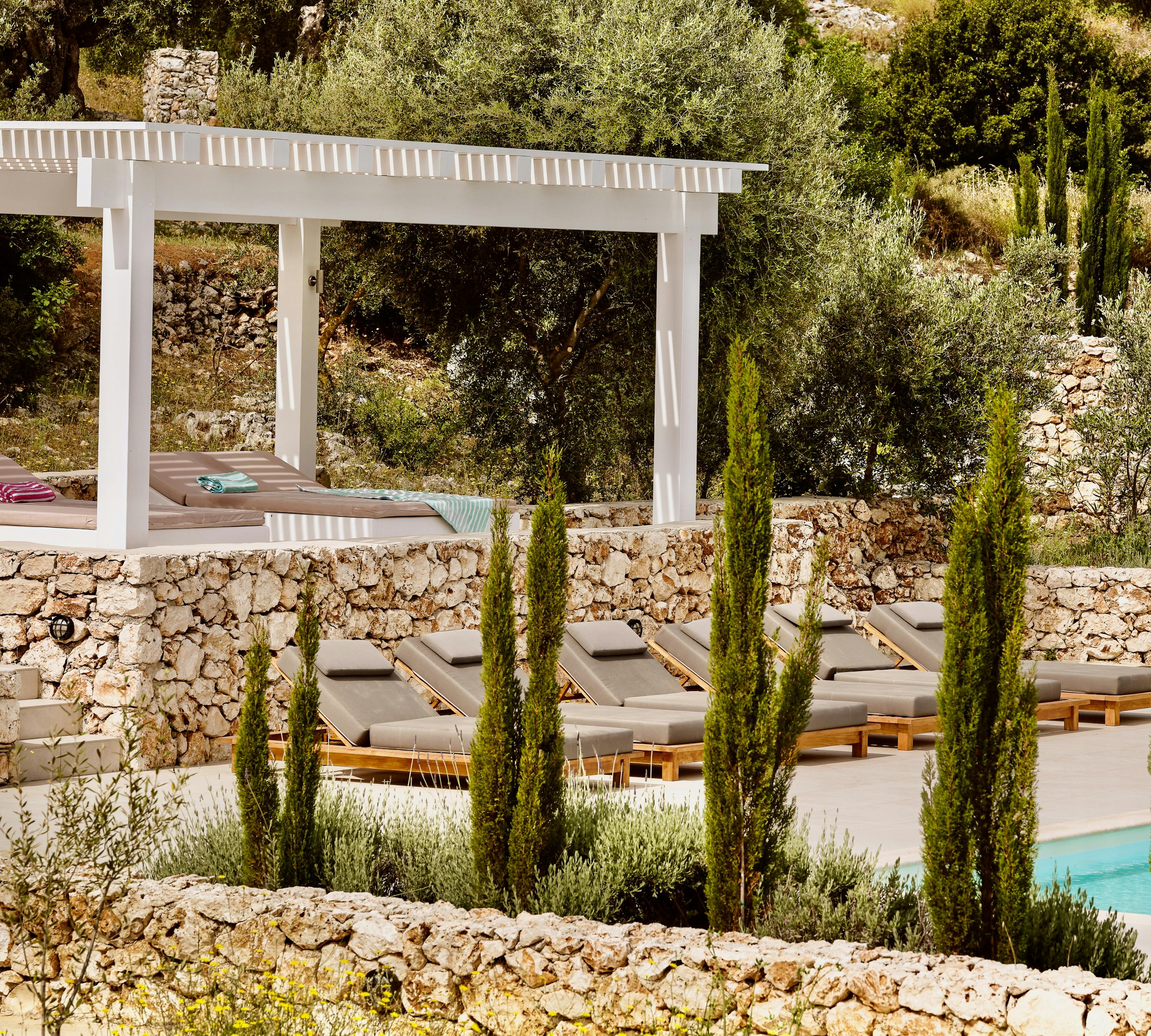 Multi-level pool deck with white pergola and wooden sun loungers with grey cushions. Tall cypress trees and stone walls define the terraced landscape design.